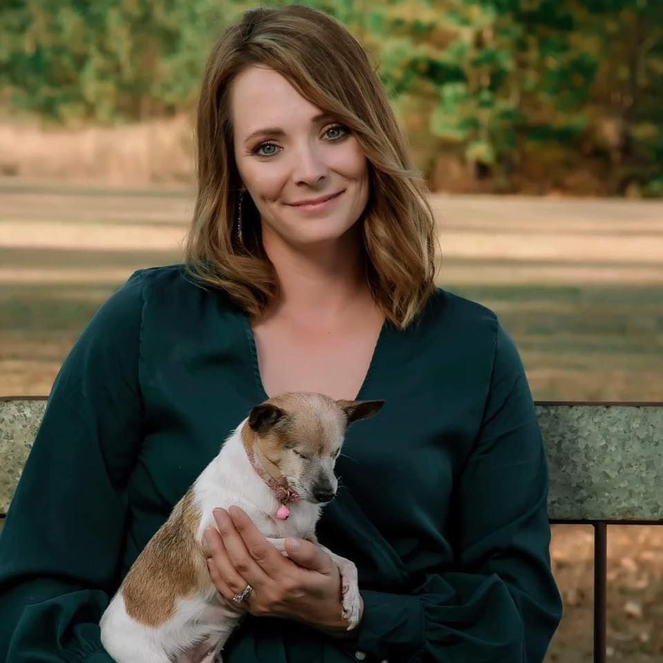 Person in emerald green sweater sitting on bench outdoors while holding small tan and white Chihuahua dog.