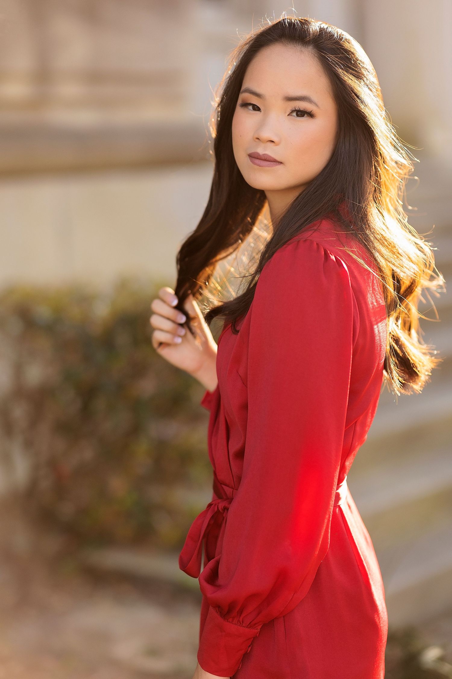 A woman in a vibrant red sweater poses outdoors during golden hour with flowing hair and natural backlighting.