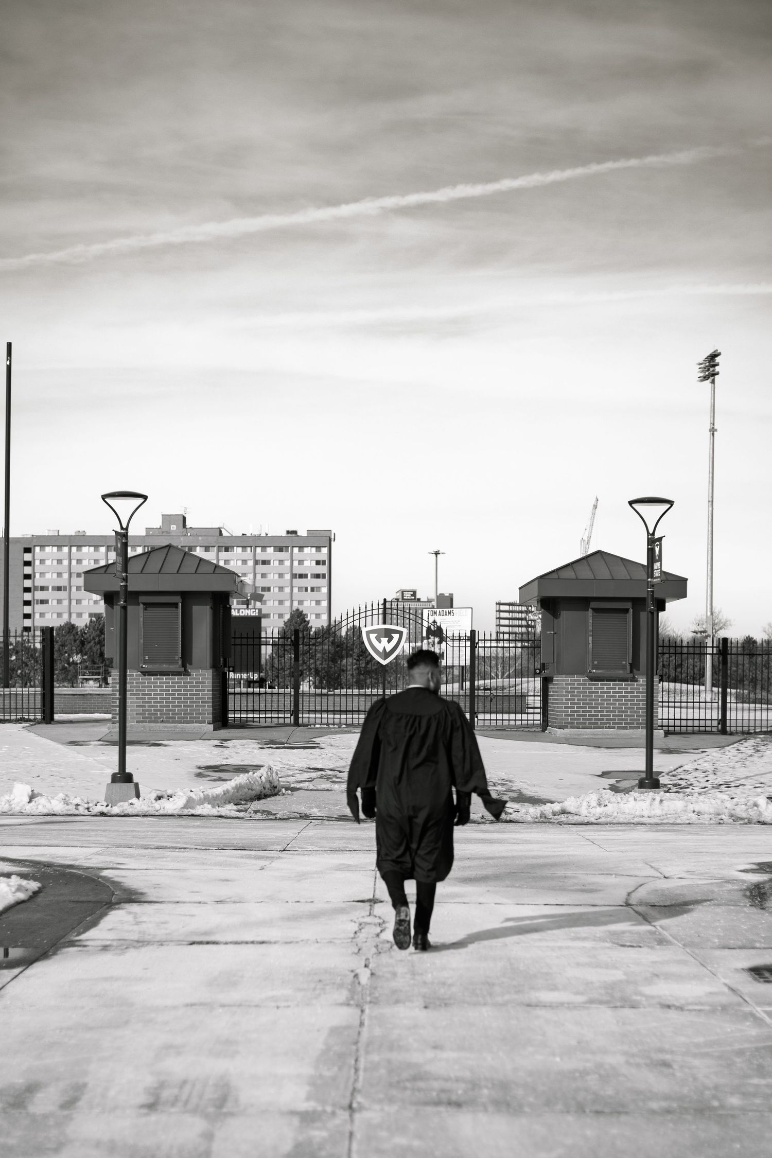 Black and white photo of a graduate in cap and gown walking toward stadium gates on a winter day.