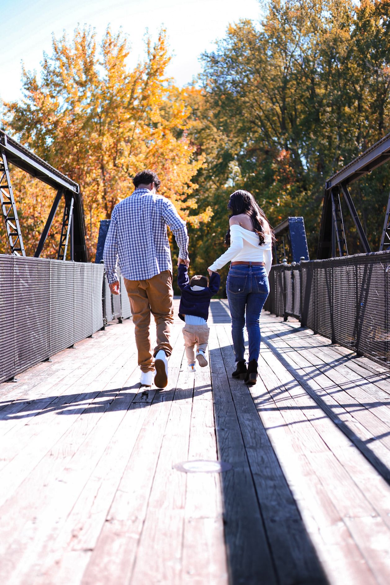 Family of three walking hand in hand across bridge in fall park.