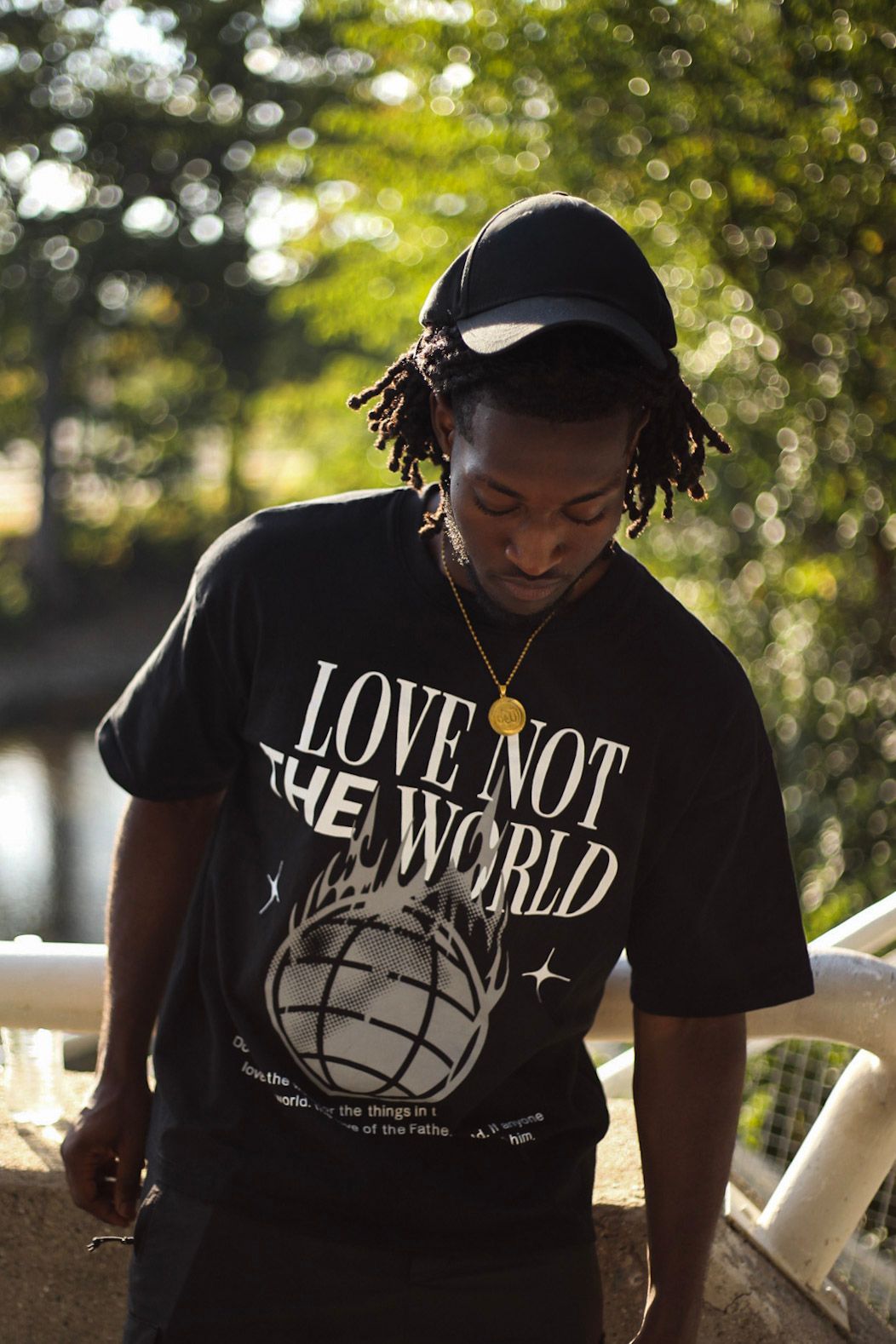 Man in black 'Love Not The World' graphic tee with gold chain and cap looking down on bridge.
