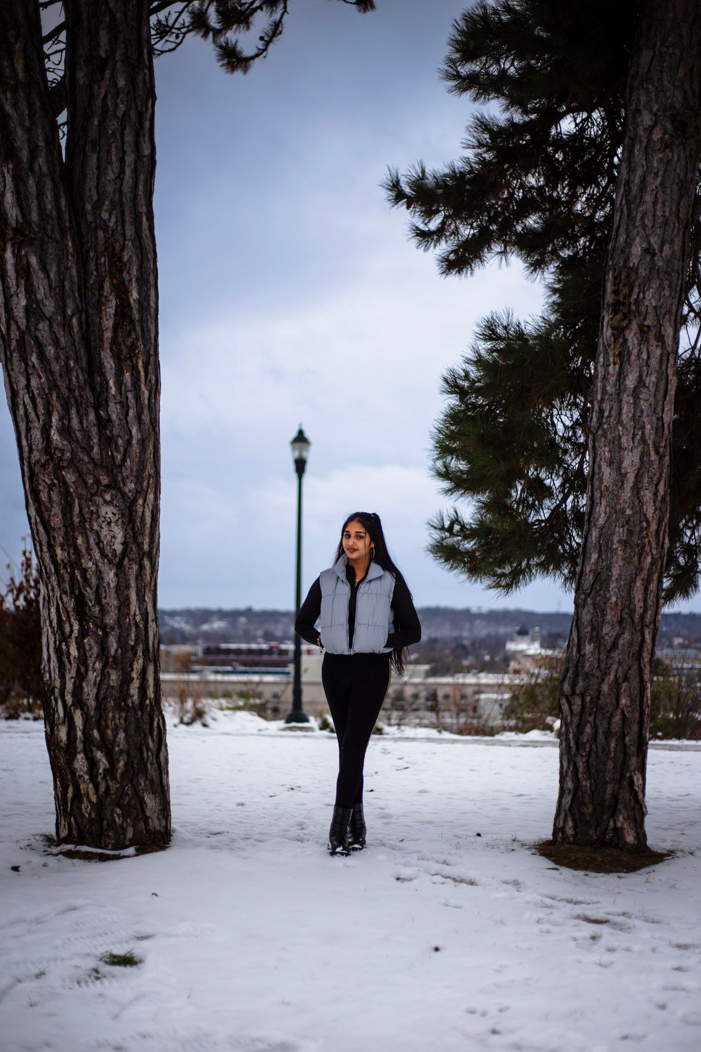 Young woman in silver vest walks between tall pine trees in a snowy urban park.