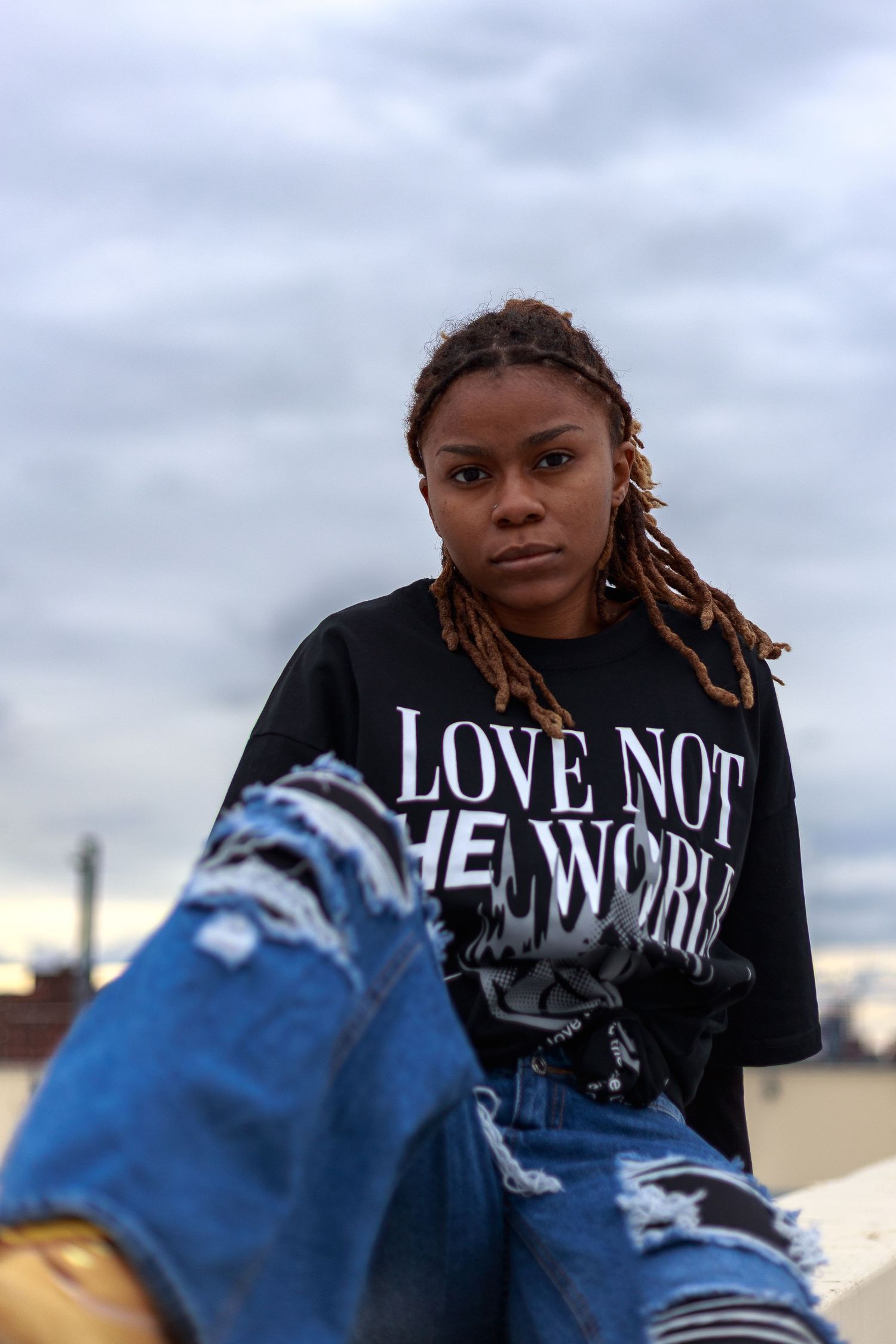 Woman in black 'Love Not The World' oversized tee and ripped jeans sits on rooftop ledge under cloudy sky.
