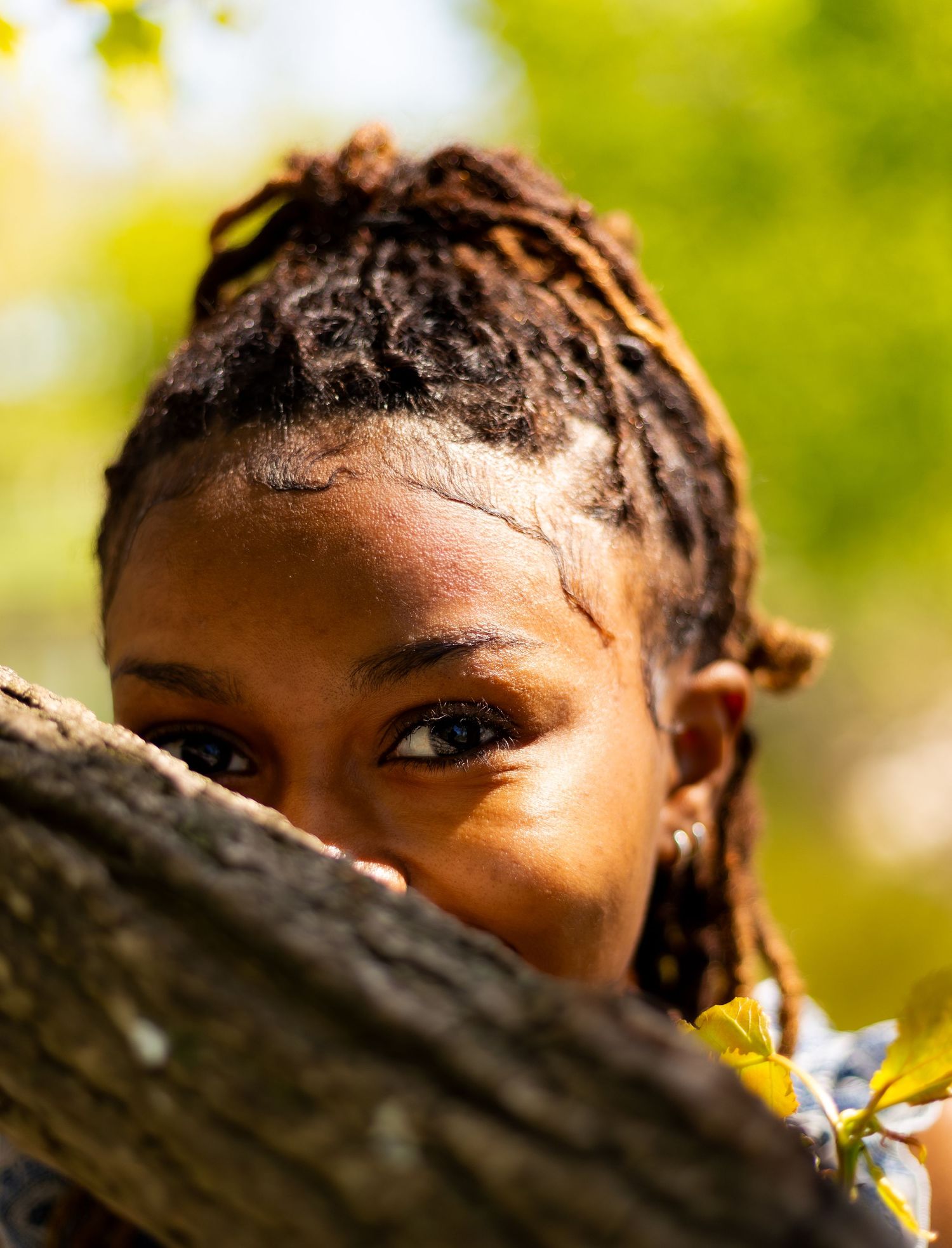 Close-up portrait of a young Black woman with locs peeking over a tree branch in a sunny green park.