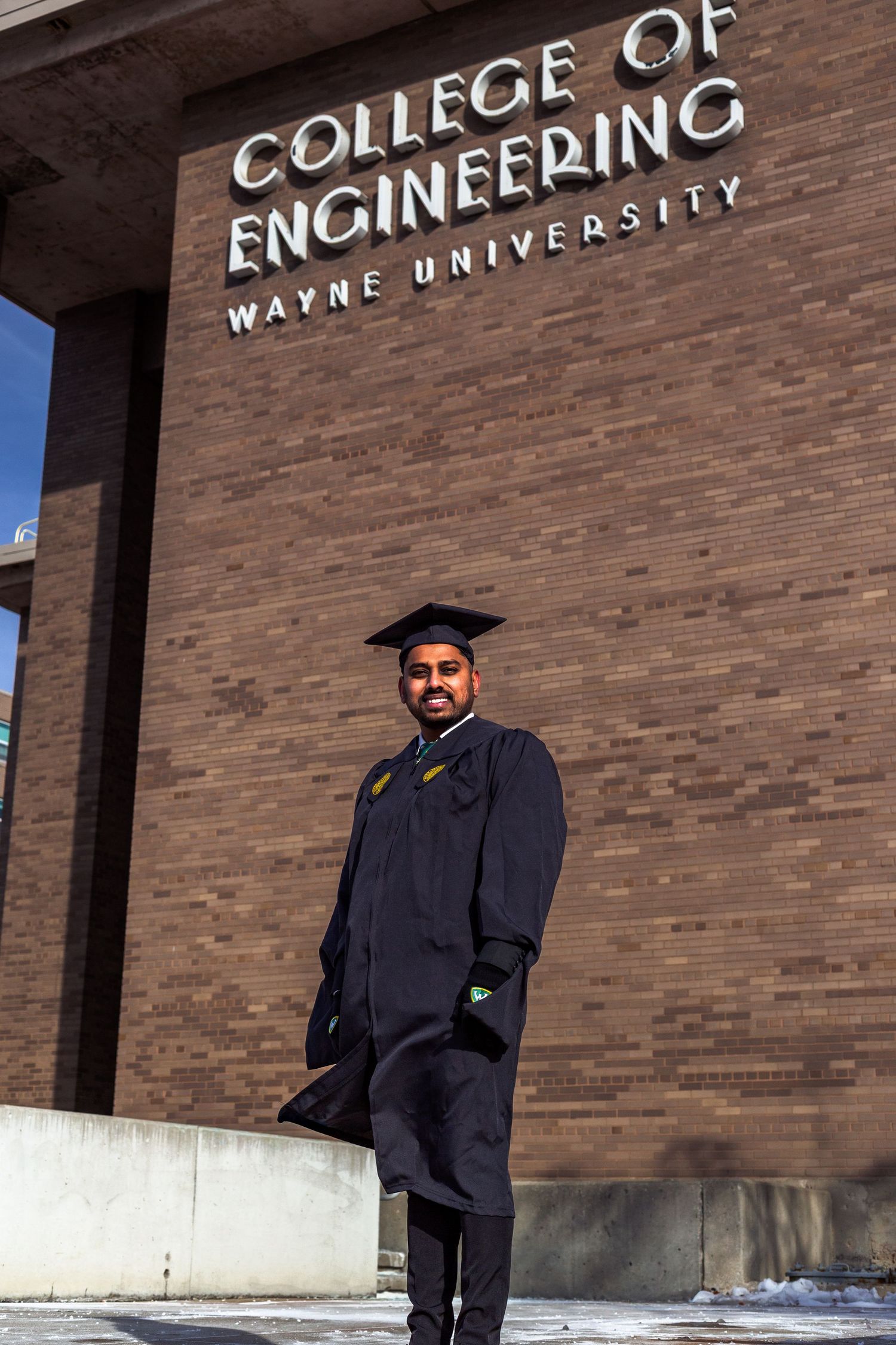 Graduate in cap and gown stands before Wayne State College of Engineering building.