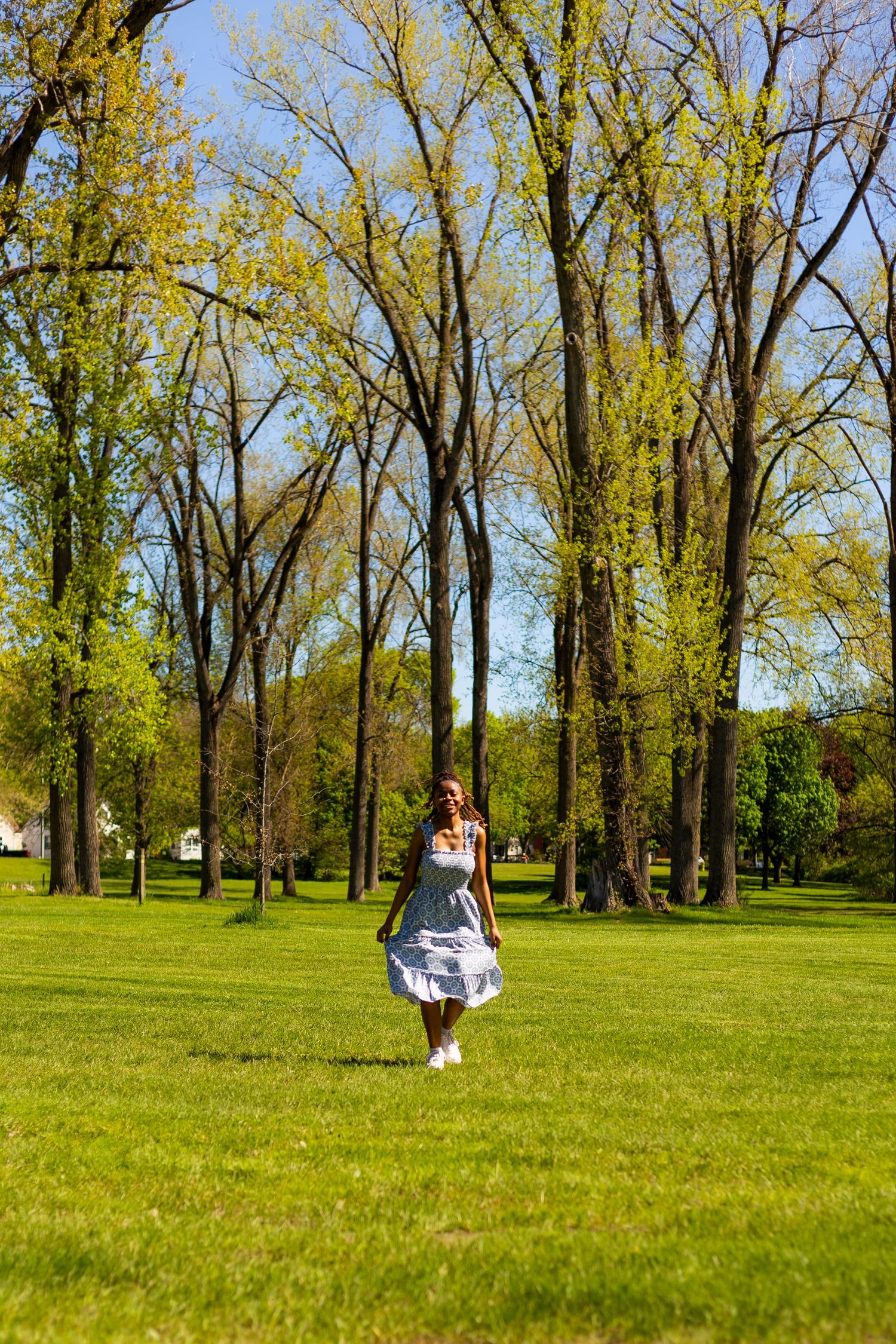 Young Black woman in a blue sundress walking across a lush green park with tall trees on a sunny day.