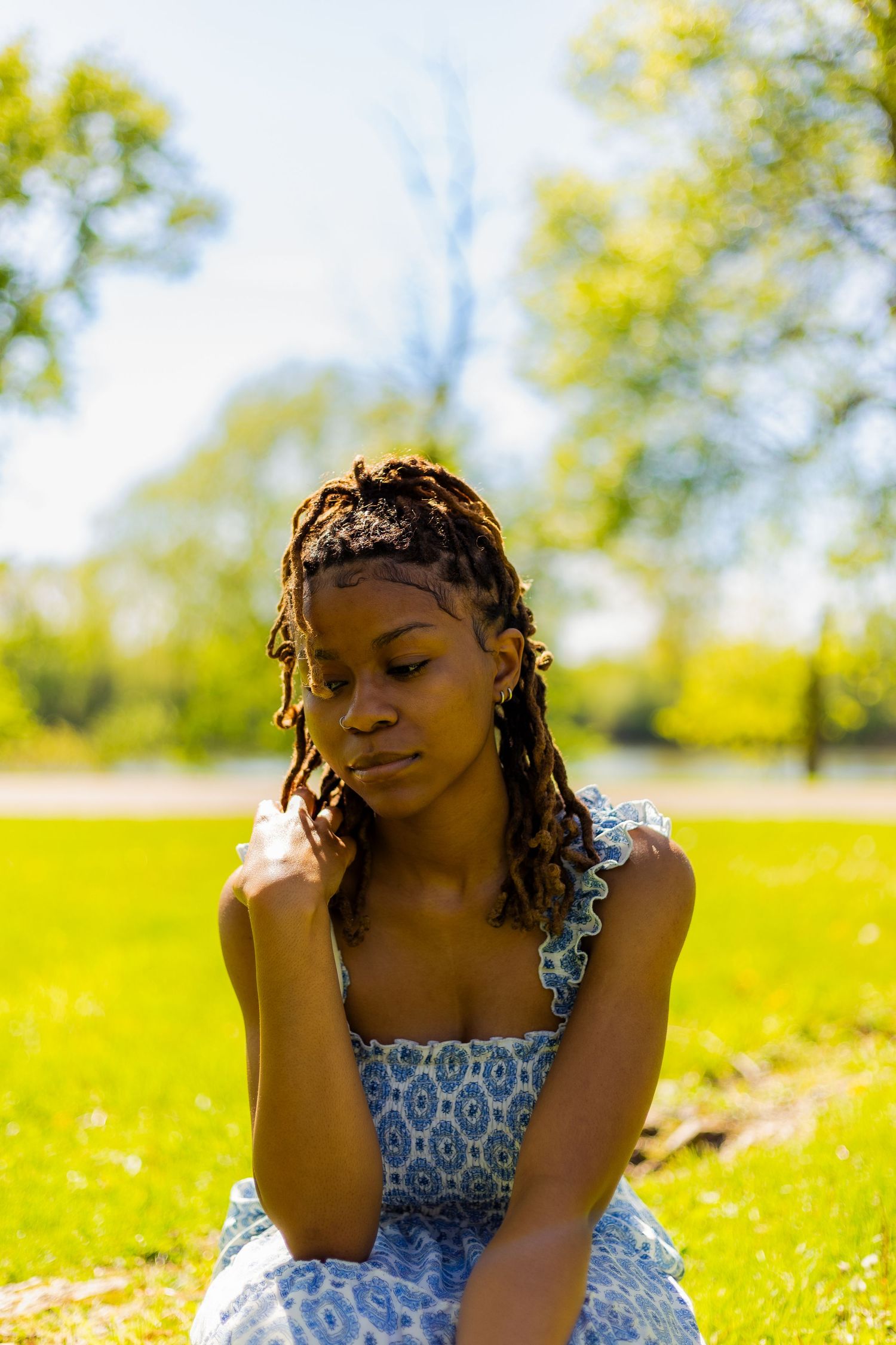 Young Black woman with braids in a blue sundress sitting on grass in a sunny park, looking down thoughtfully.