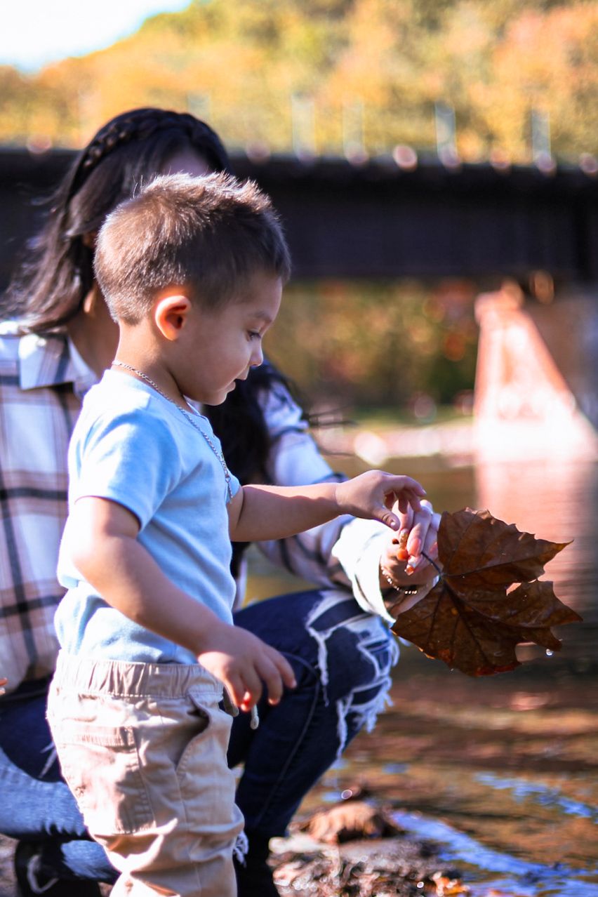 A young boy examines a large autumn leaf near a river while his mother sits close behind him.