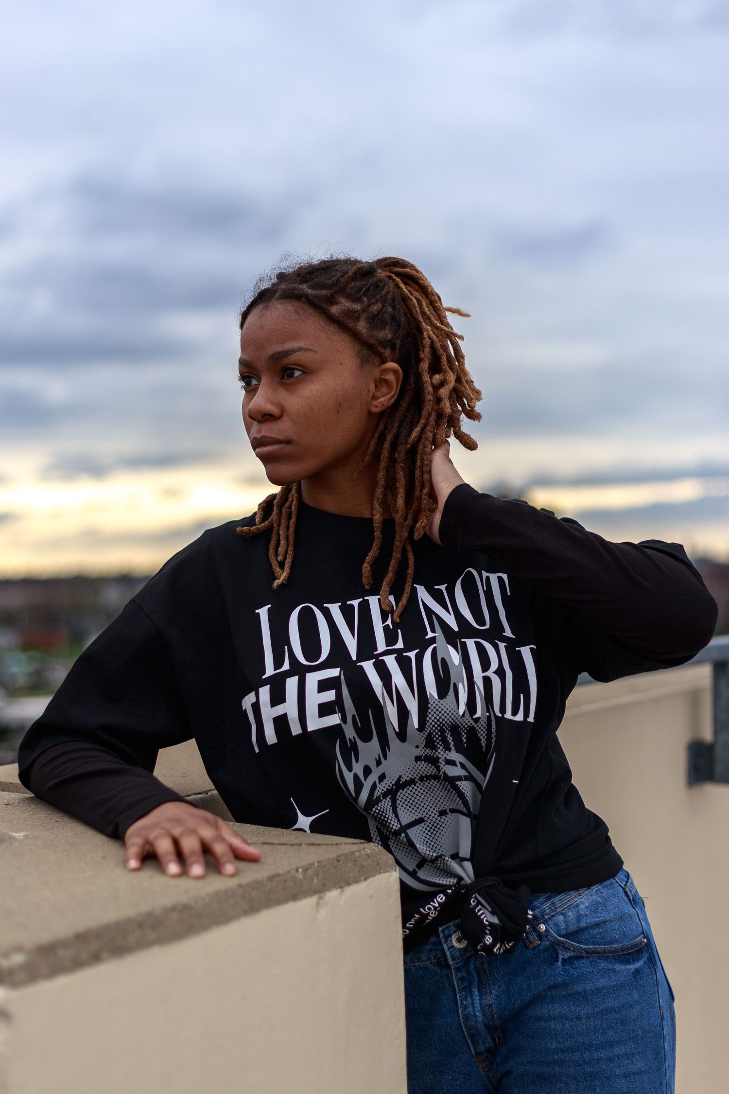 Young woman with locs wearing a black Love Not The World sweatshirt leans on a rooftop wall at sunset.
