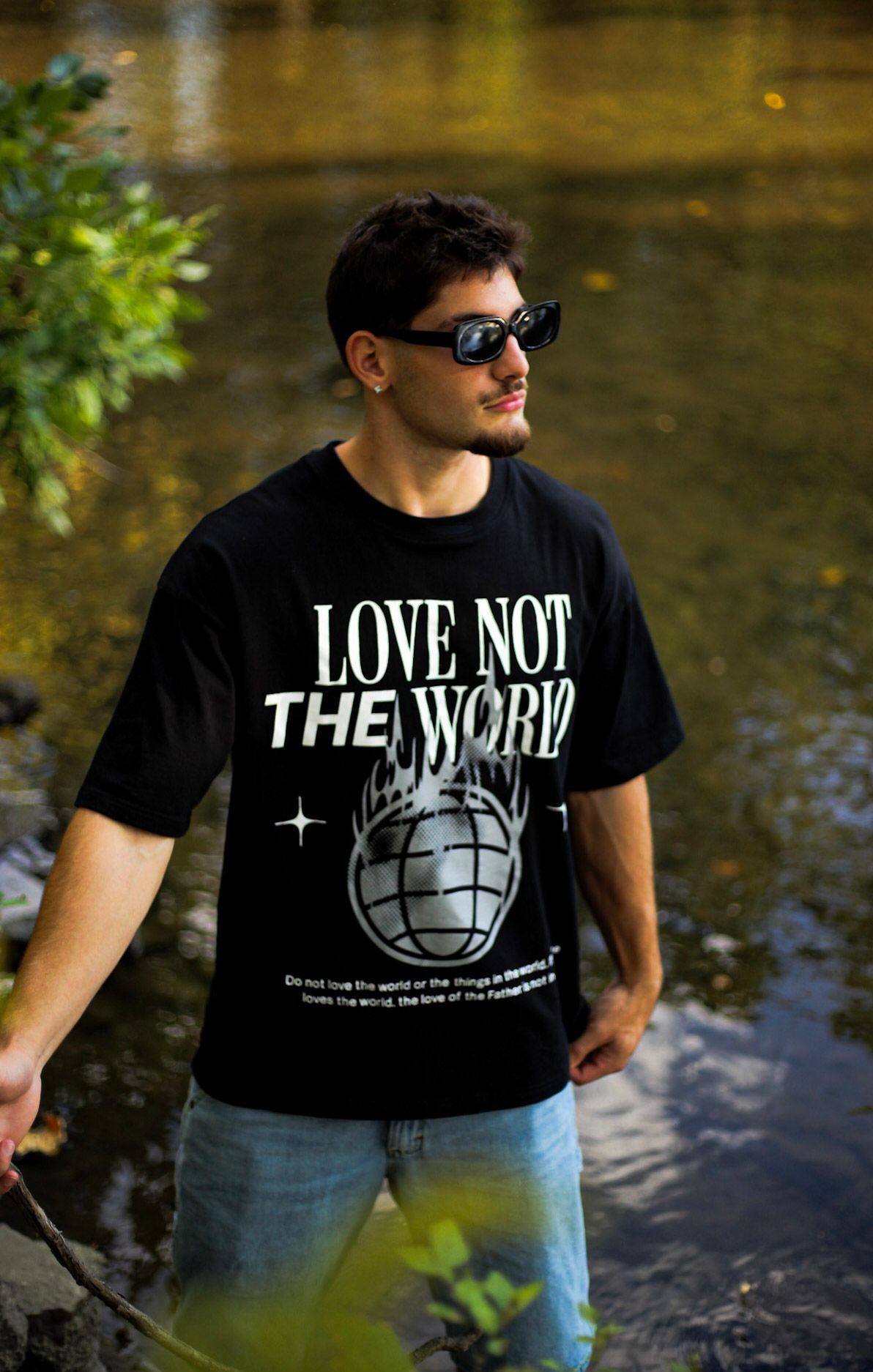 Man in black 'Love Not The World' graphic tee with sunglasses standing near reflective water outdoors.