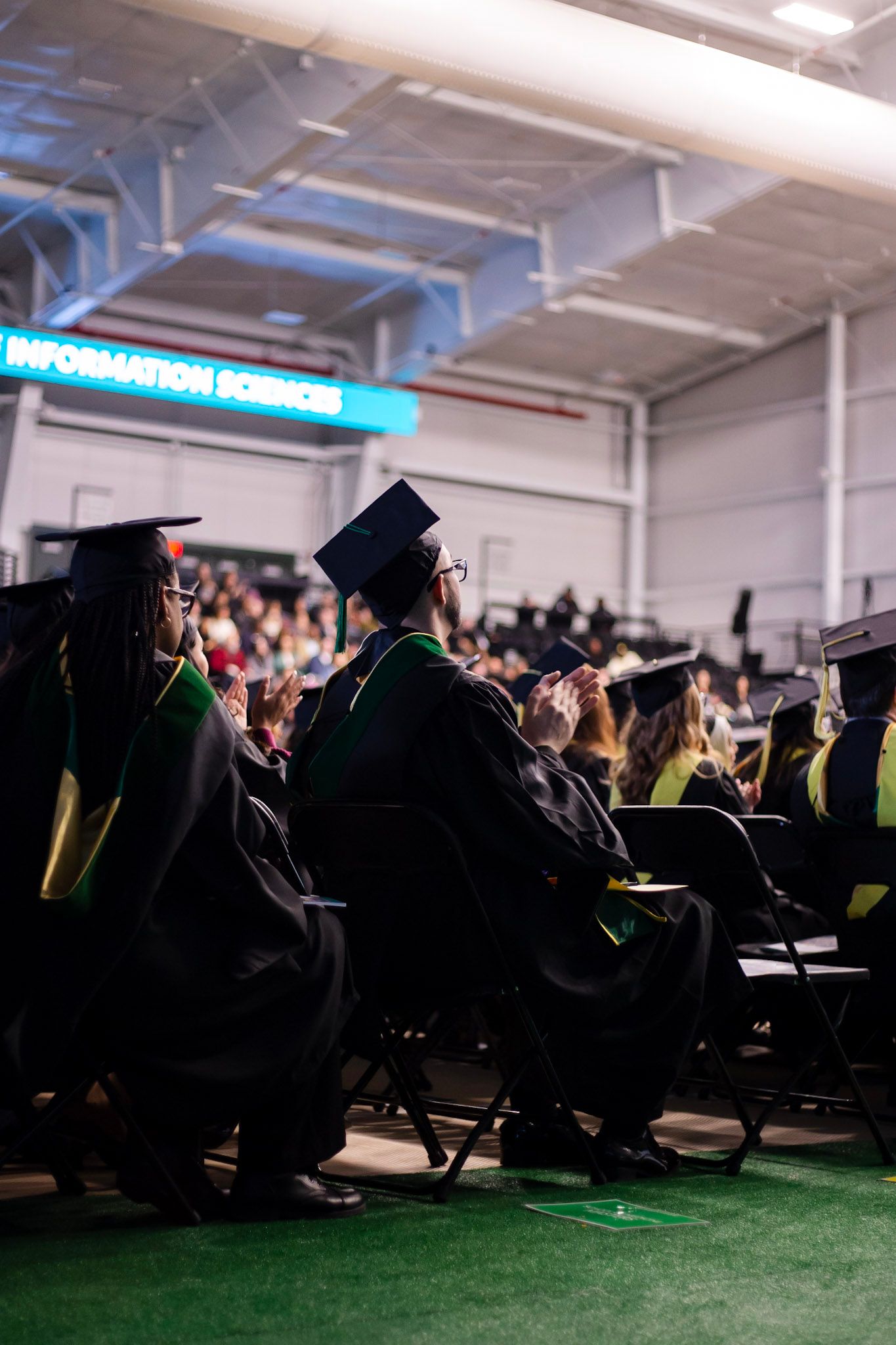 Graduates in black caps and gowns seated at commencement with Information Sciences banner glowing in the background.