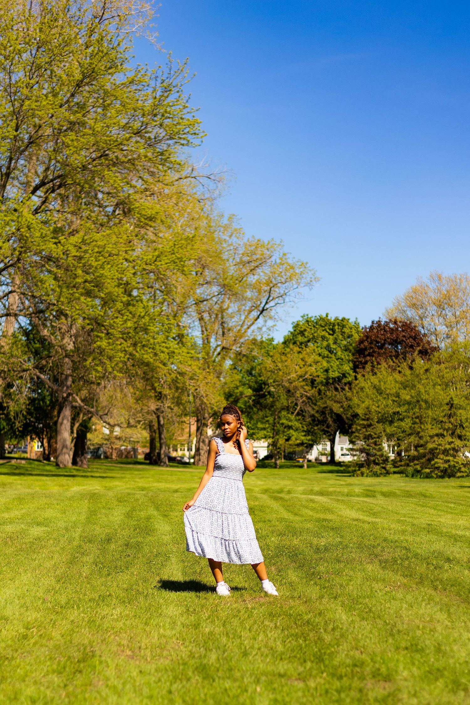 Young Black woman in blue floral sundress stands on green lawn surrounded by spring trees.