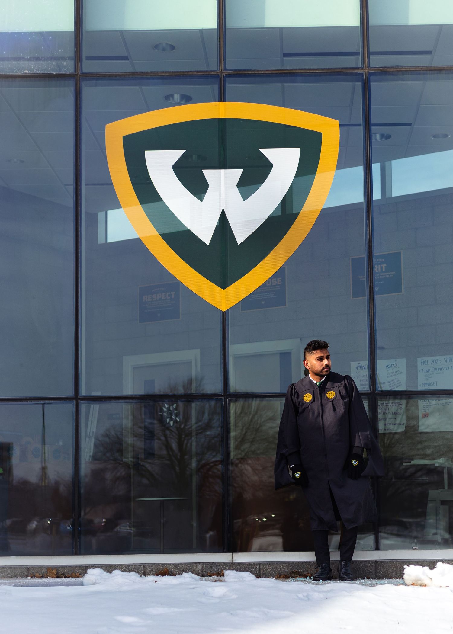 Wayne State University graduate standing in snow before large W shield logo on glass building.