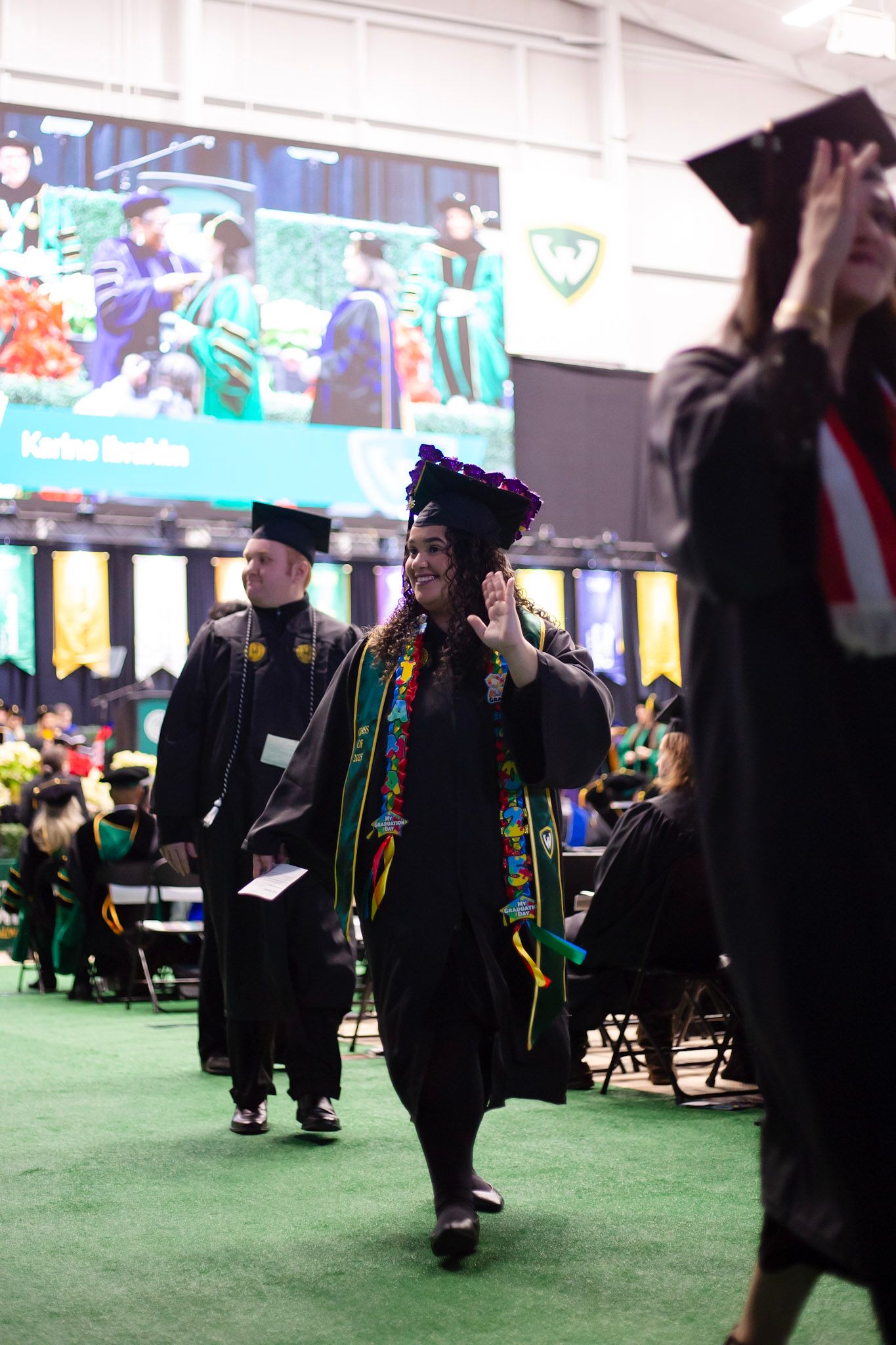 Decorated graduate waves while walking across green at university commencement ceremony.