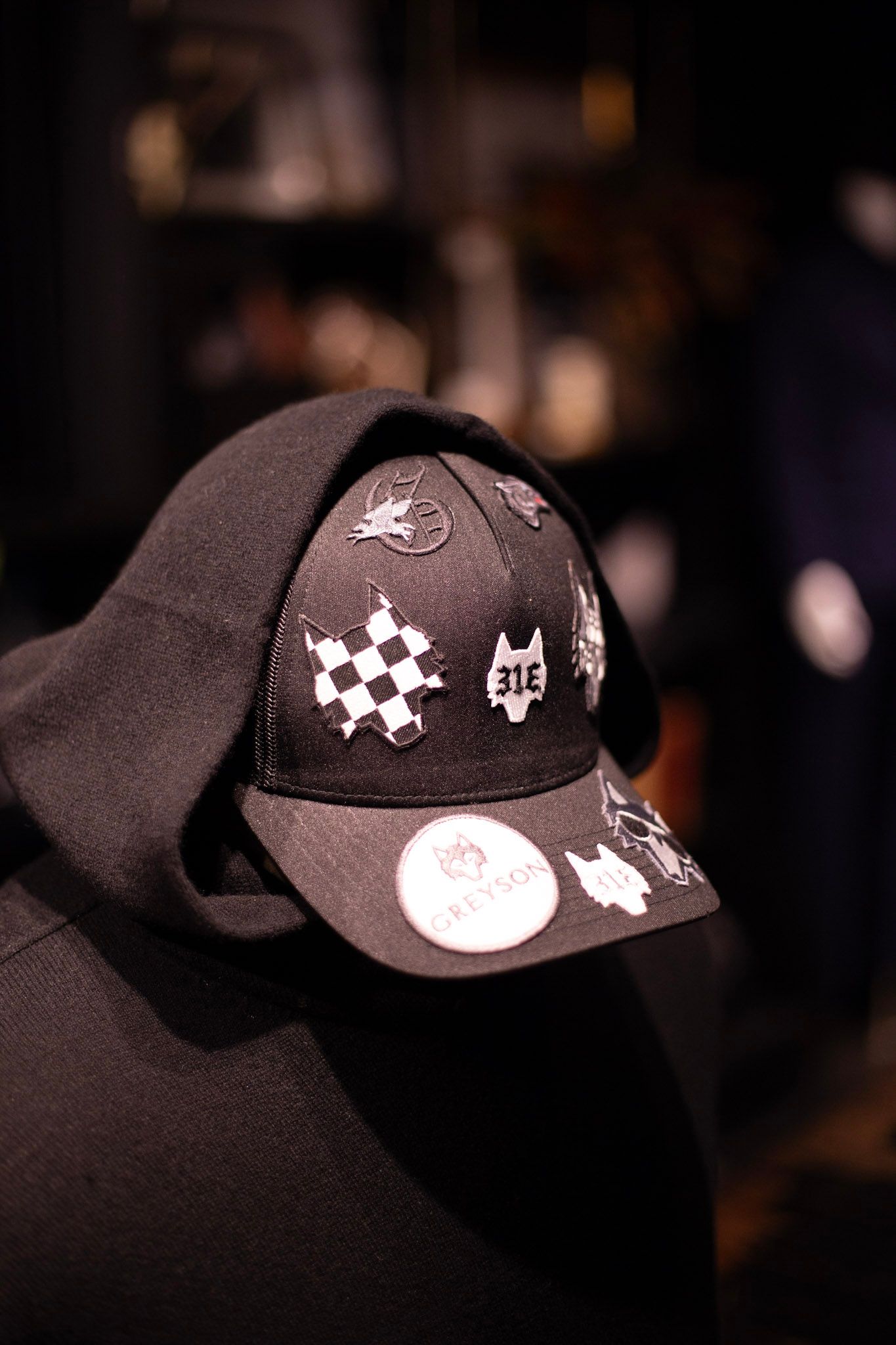 Black baseball cap with white embroidered patches and pins displayed on a hooded mannequin in a dark setting.