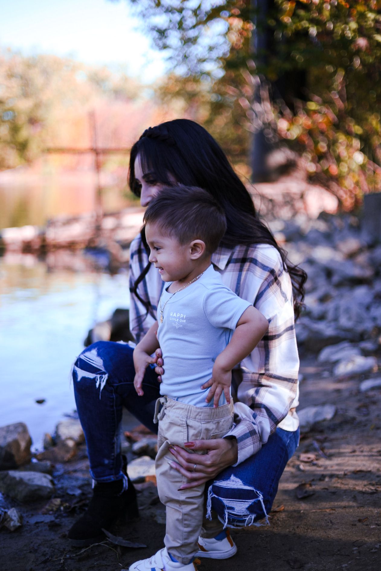 A mother in a plaid shirt kneels beside her young toddler near a rocky riverbank in fall.