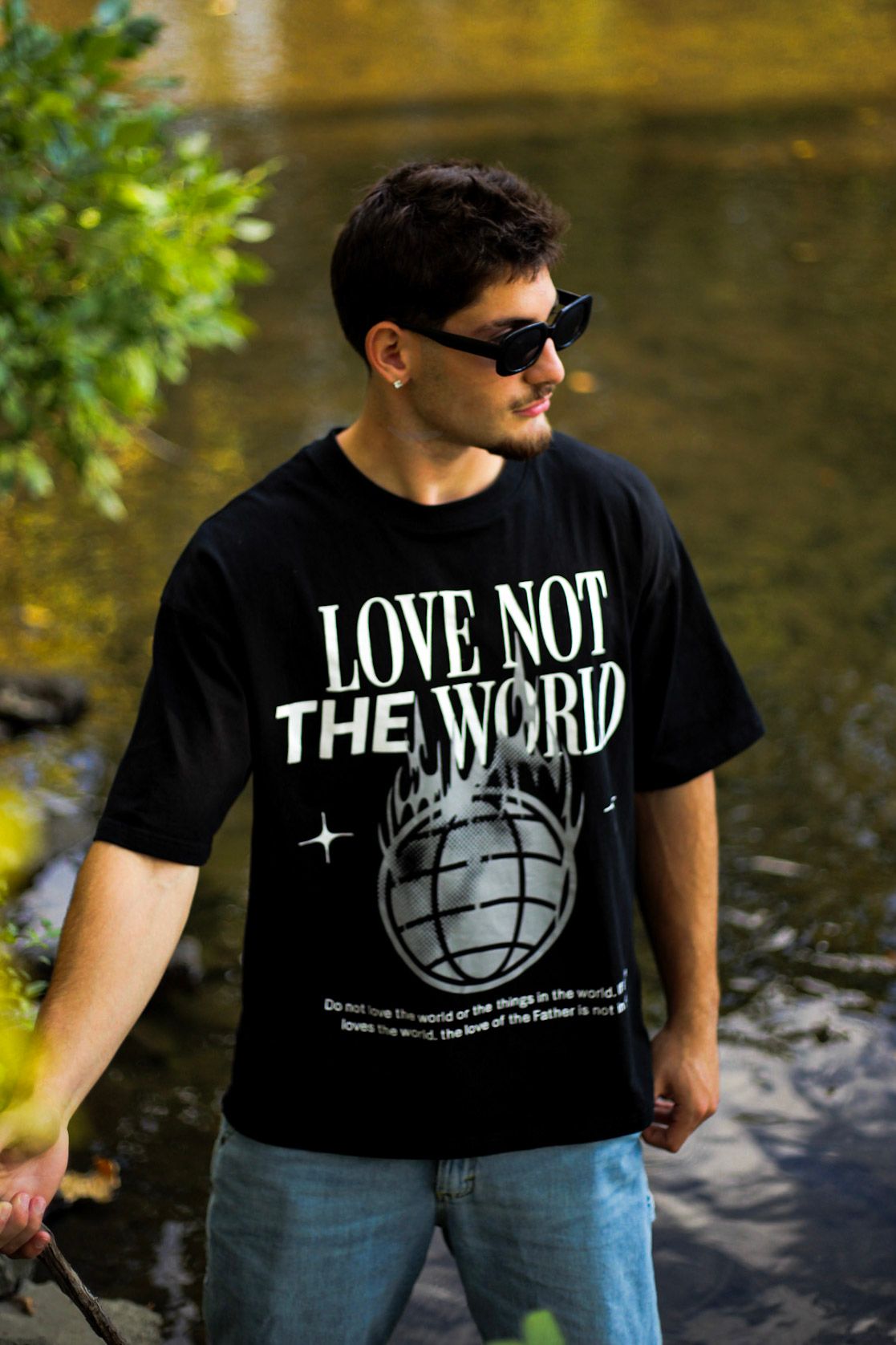Young man in sunglasses wearing black Love Not The World oversized tee standing by a river.