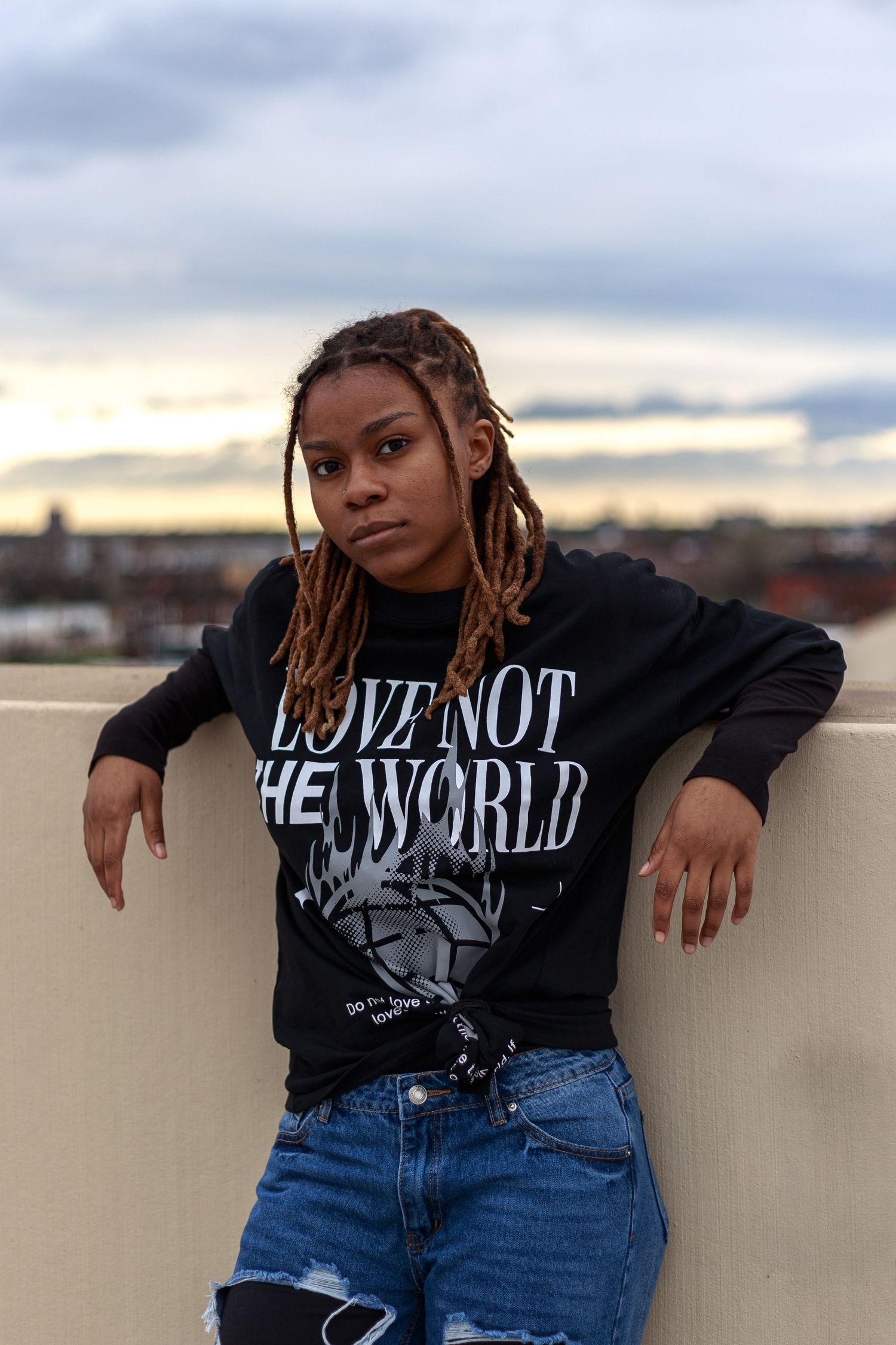 Woman with blonde locs in a black Love Not The World sweatshirt leans against a wall at golden hour.