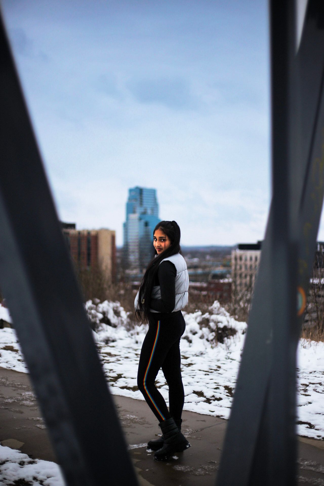 Young woman in silver vest and black pants smiles near urban snowy cityscape background.