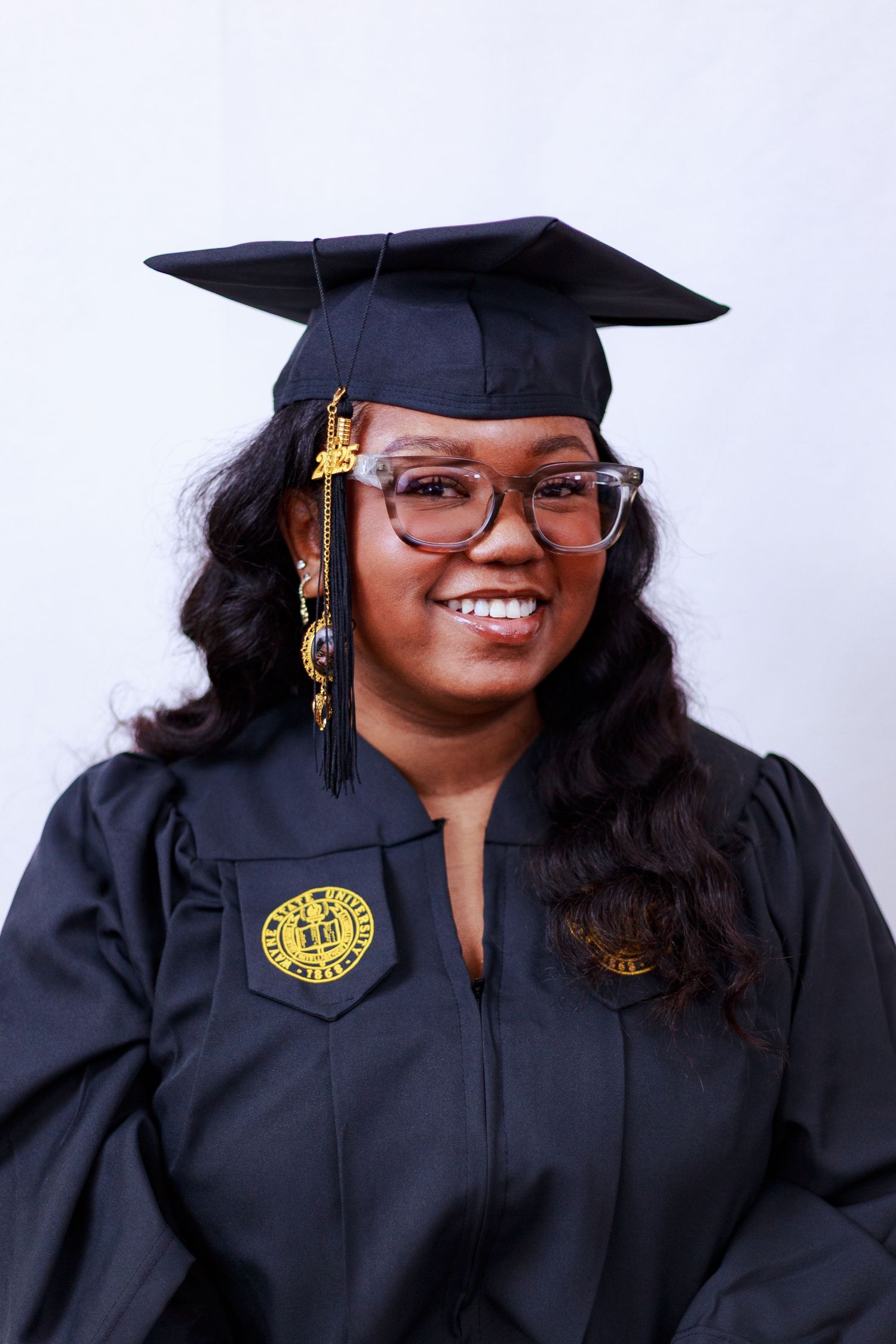 Smiling young woman in black graduation cap and gown with gold tassel and emblem against white background.