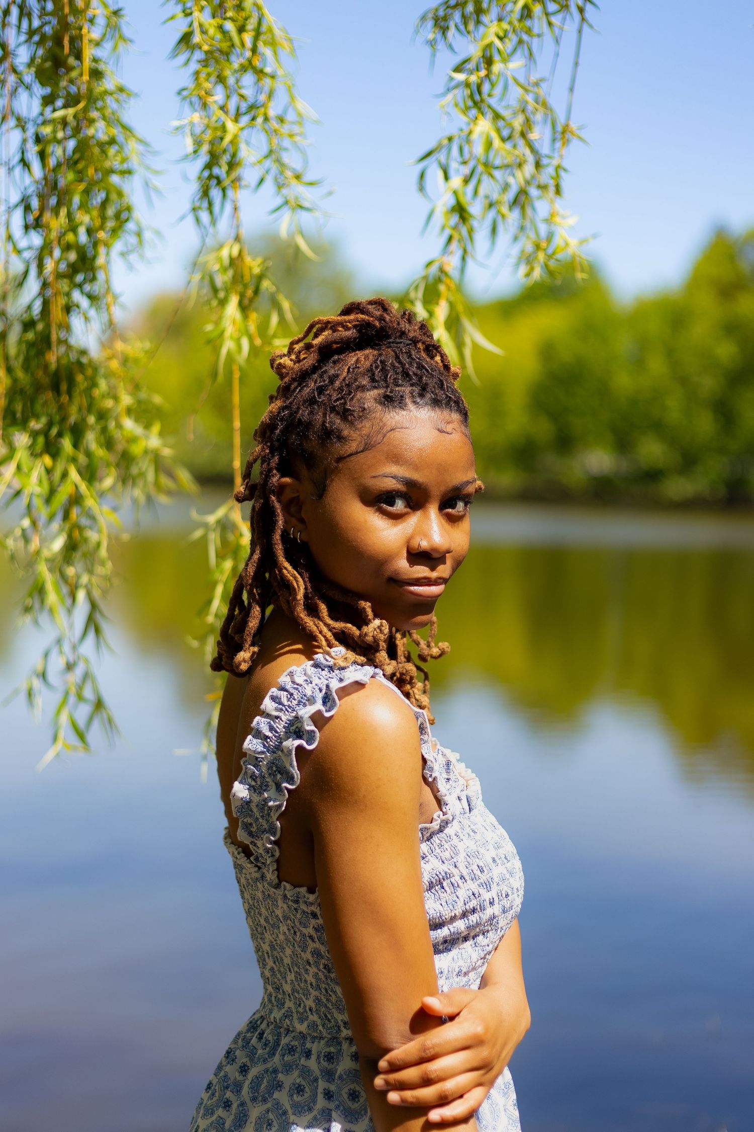 Young Black woman with dreadlocks in blue sundress poses by lake under weeping willow branches.