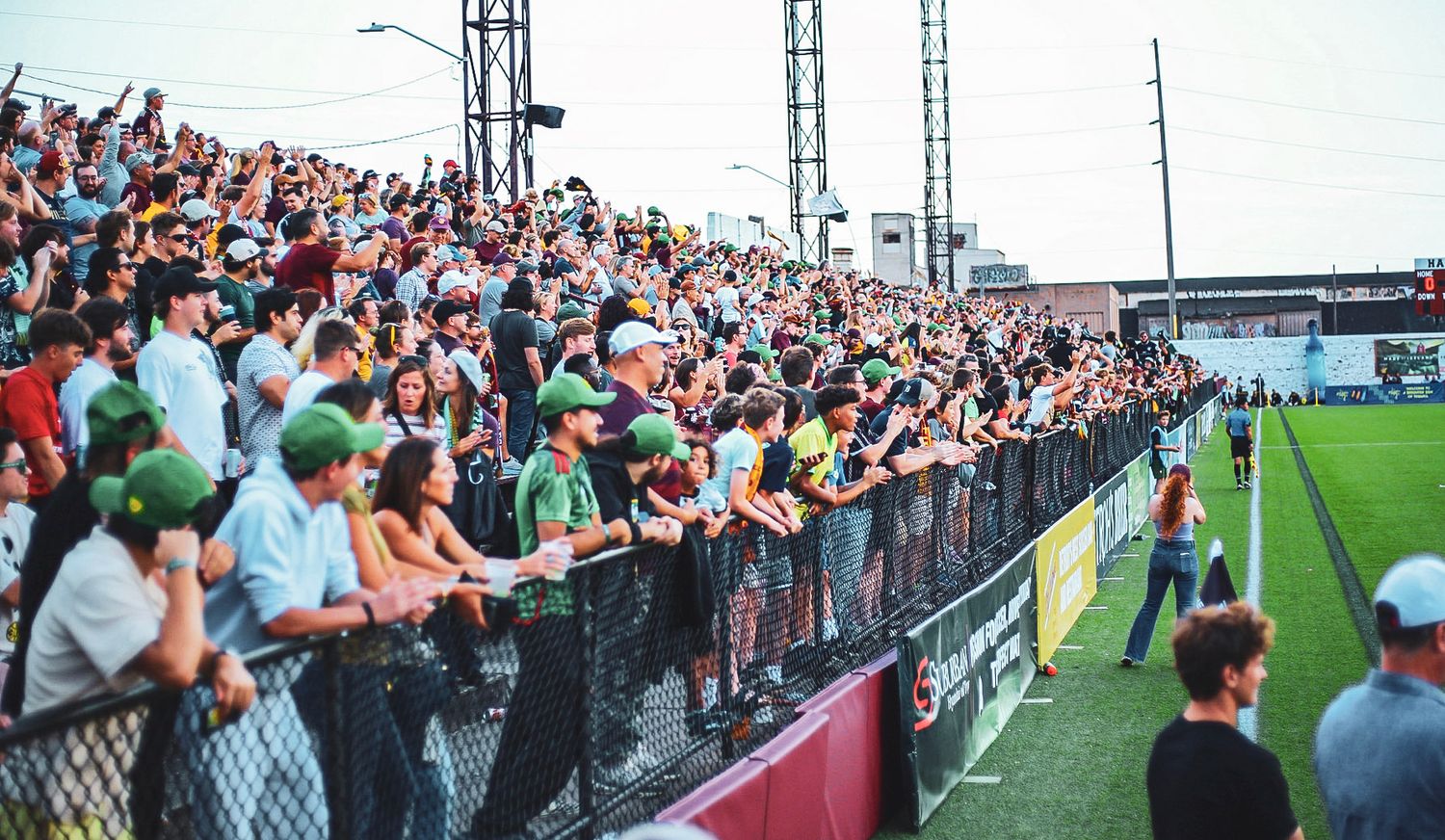 Enthusiastic crowd of fans in green hats pressed against the barrier at a soccer stadium during a lively match.