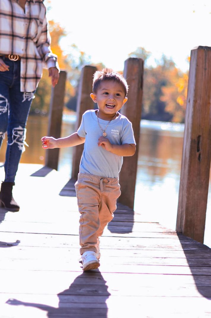 Happy toddler boy running on wooden dock near lake with fall foliage in background.