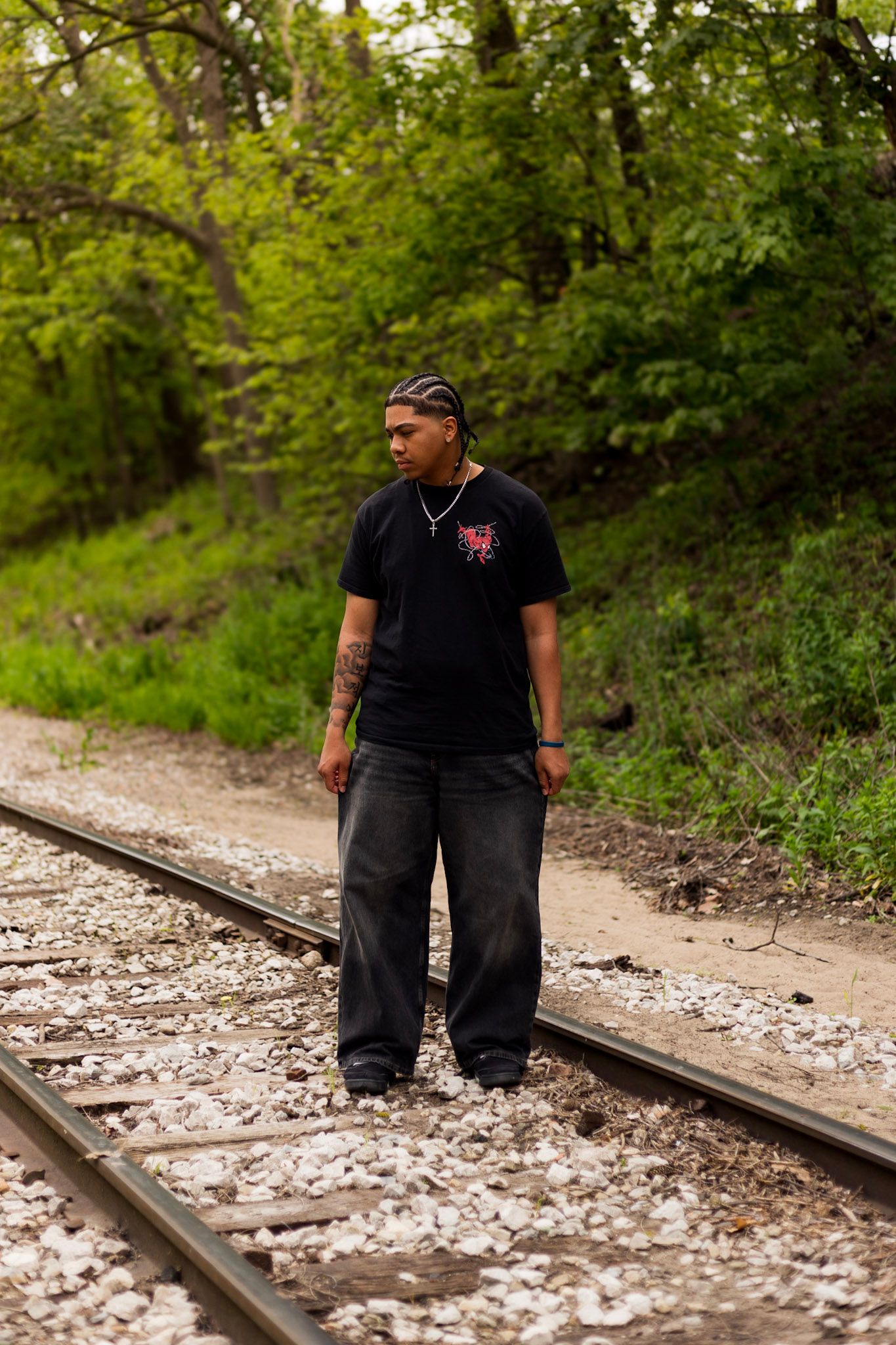 Young tattooed man in black tee and baggy jeans stands on railroad tracks surrounded by trees.