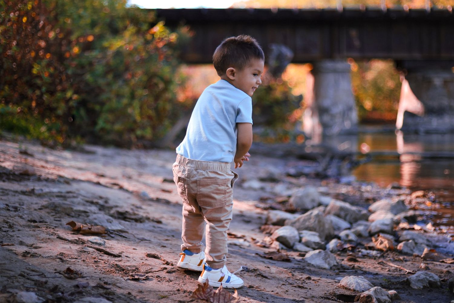Toddler boy in white shirt and tan pants standing on rocky shore near bridge.