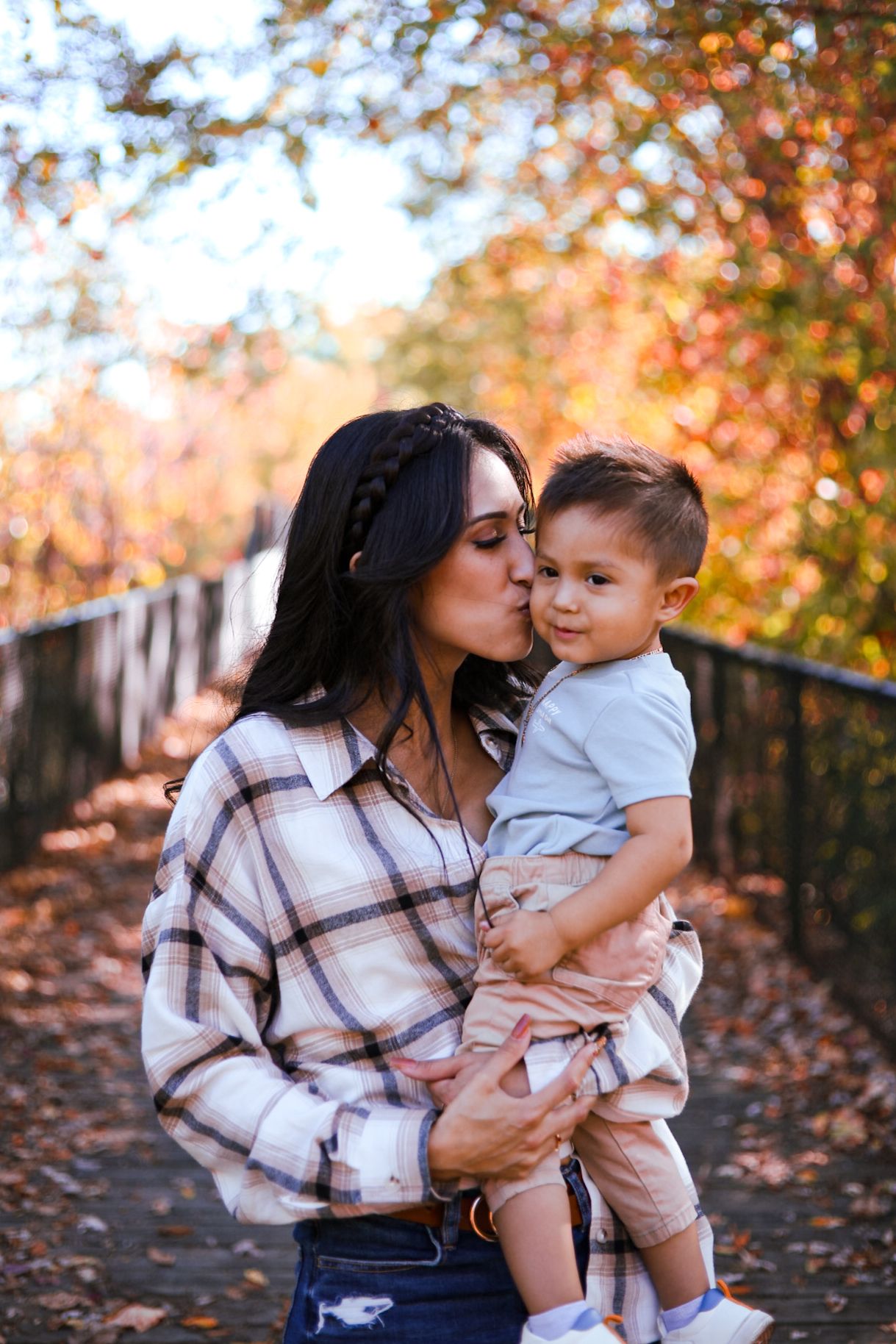 Loving mother kissing her young toddler son on a wooden bridge surrounded by fall leaves.