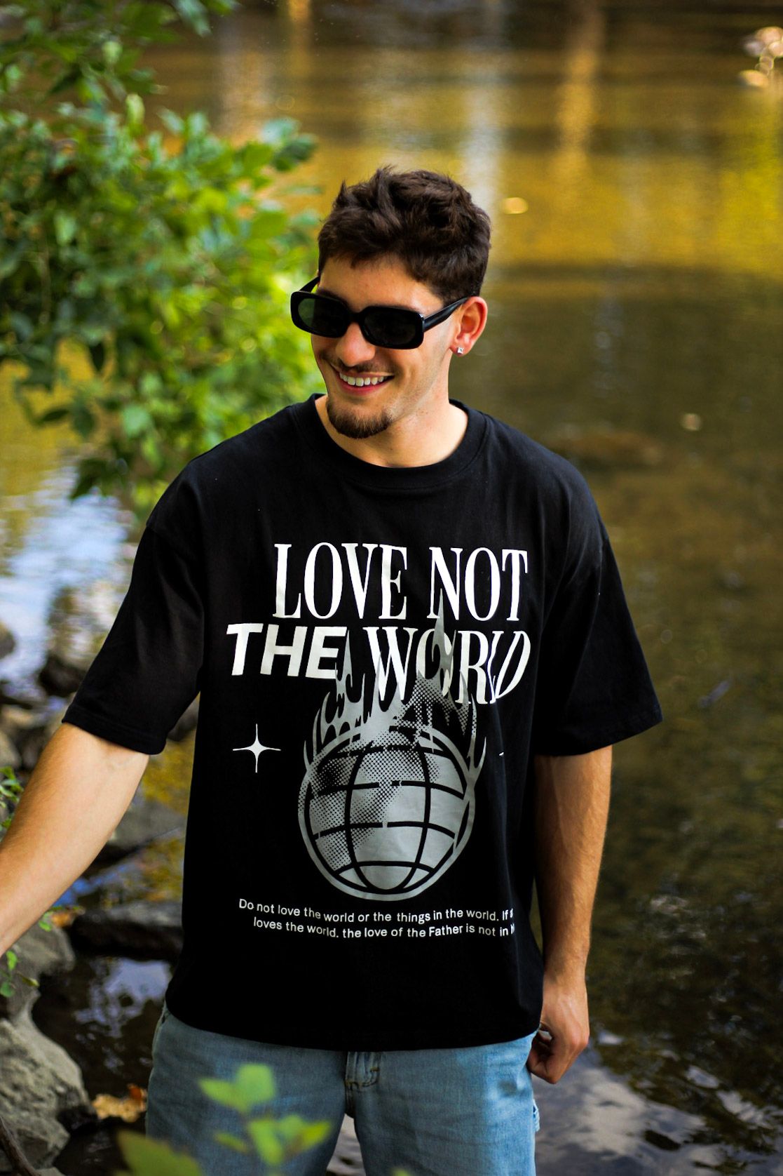 Smiling man in sunglasses wearing black Love Not The World oversized graphic tee outdoors.