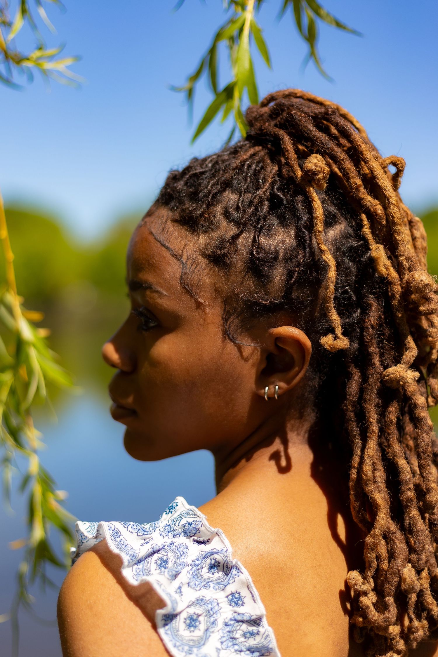 Side profile of young Black woman with long brown dreadlocks in blue dress near sunlit lake.