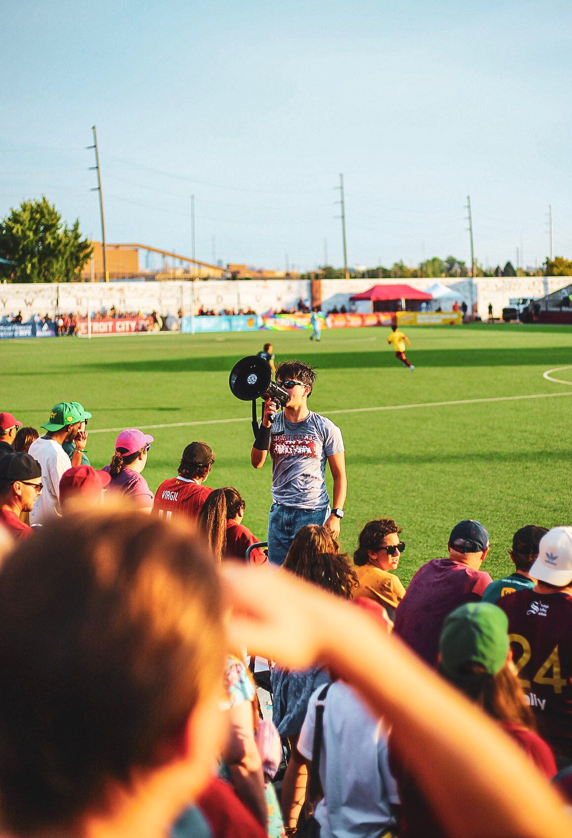 A cameraman films Detroit City FC soccer match while fans in maroon jerseys cheer from the sidelines.
