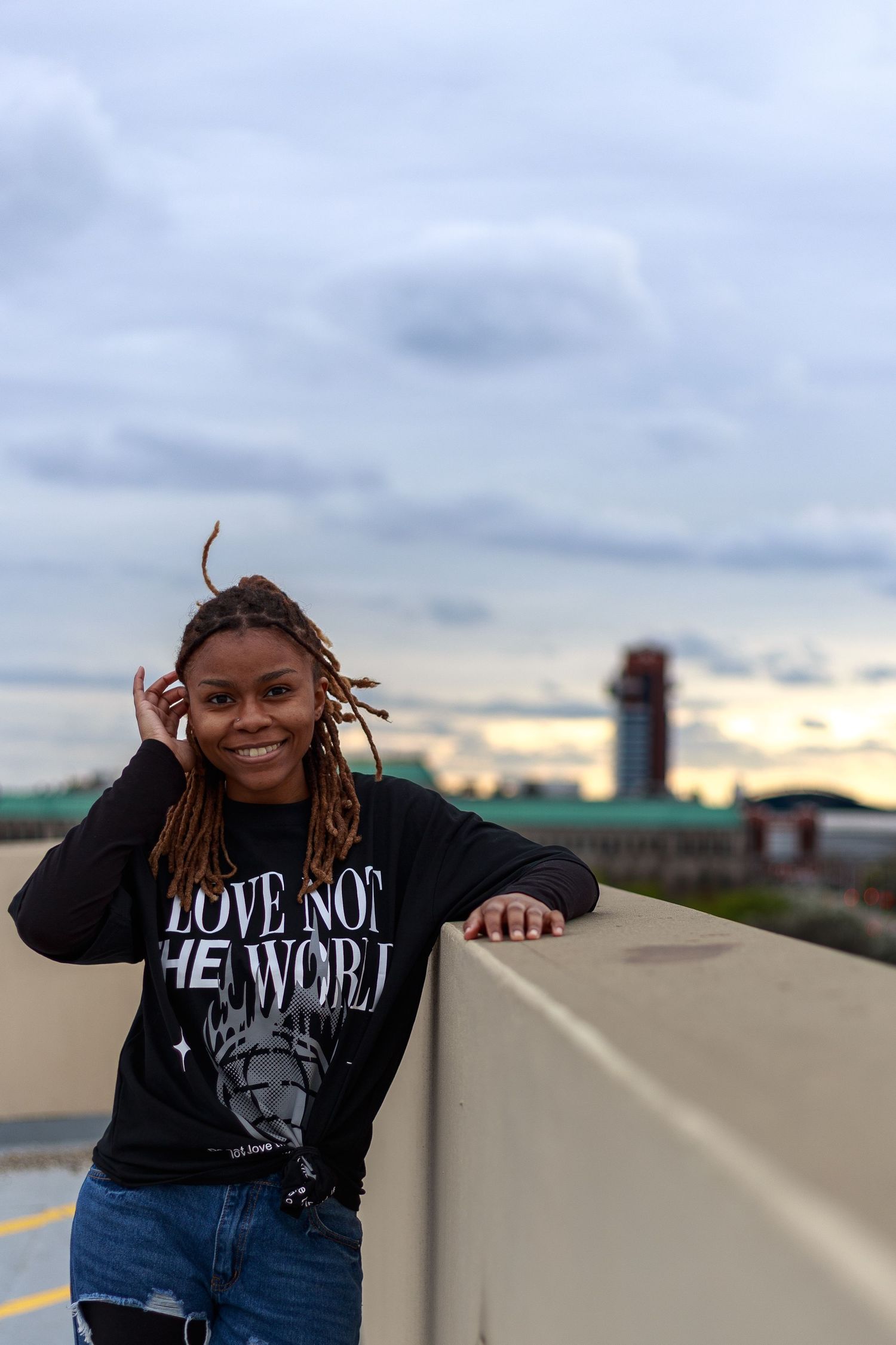 Smiling woman with locs wearing a Love Not The World black sweatshirt poses on a rooftop at sunset.