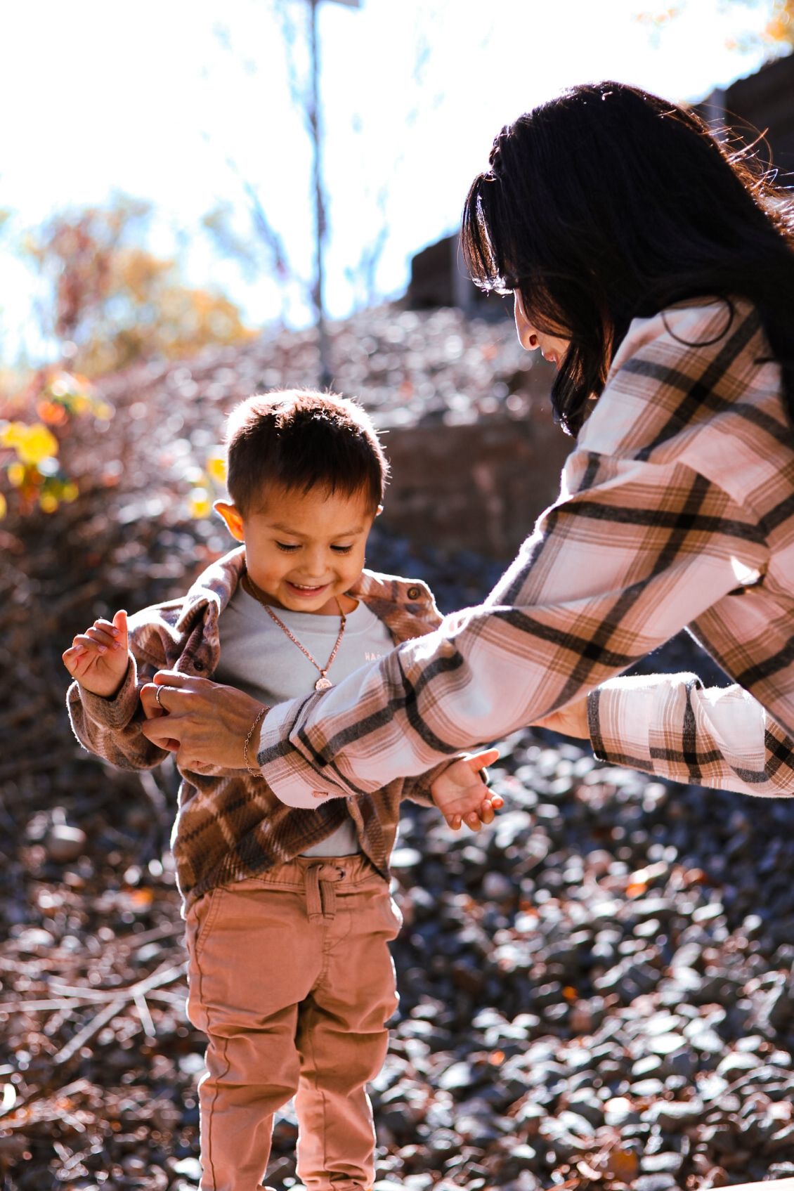 A mother in a plaid coat joyfully holds her laughing toddler son outdoors in autumn leaves.