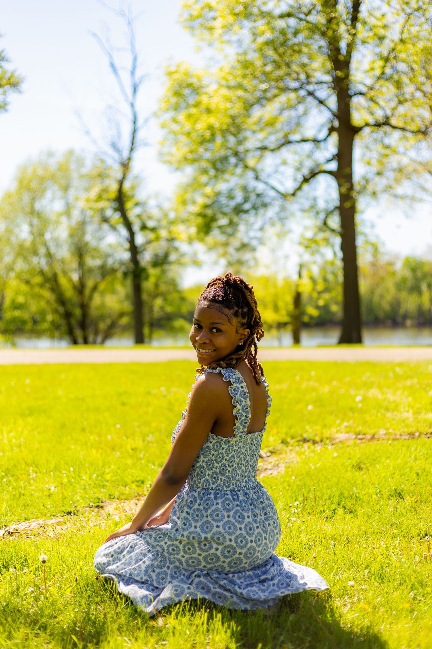 Young Black woman in a blue floral dress sitting on green grass in a park, smiling over her shoulder.