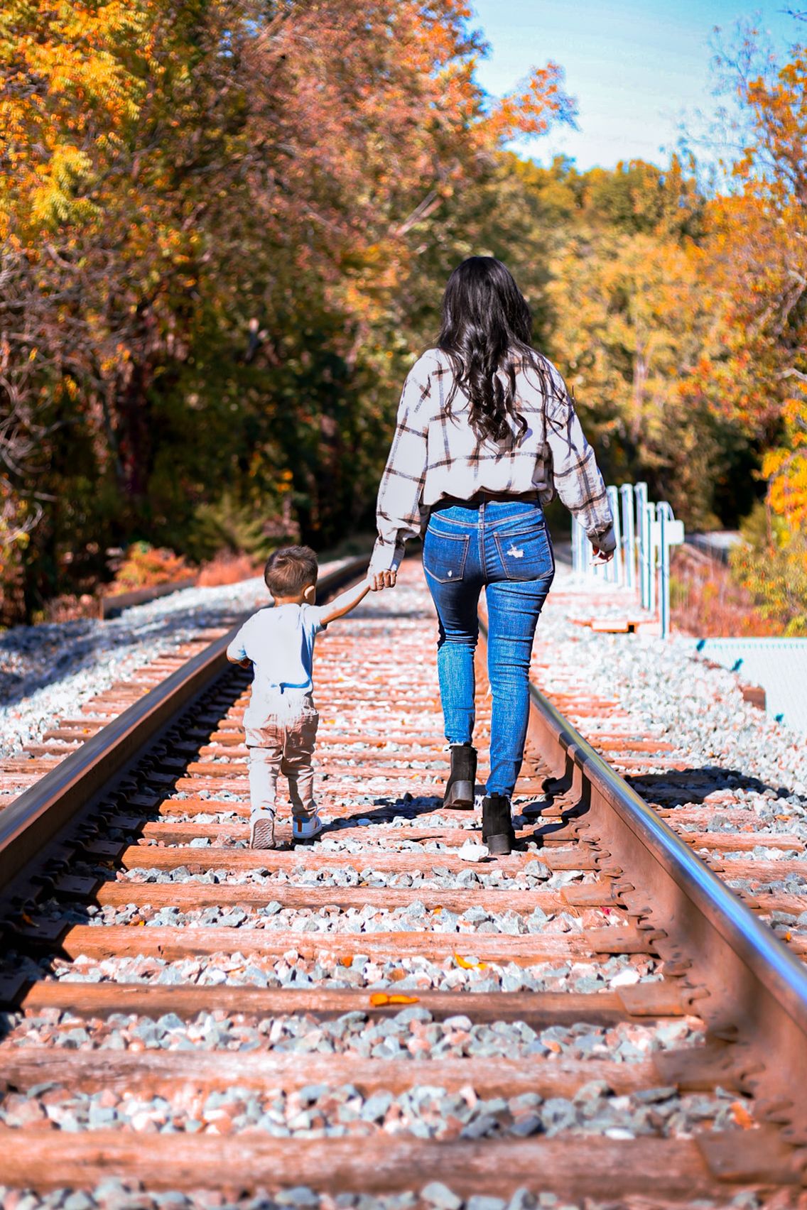 Mother and toddler son walking hand in hand along rustic railroad tracks in autumn.
