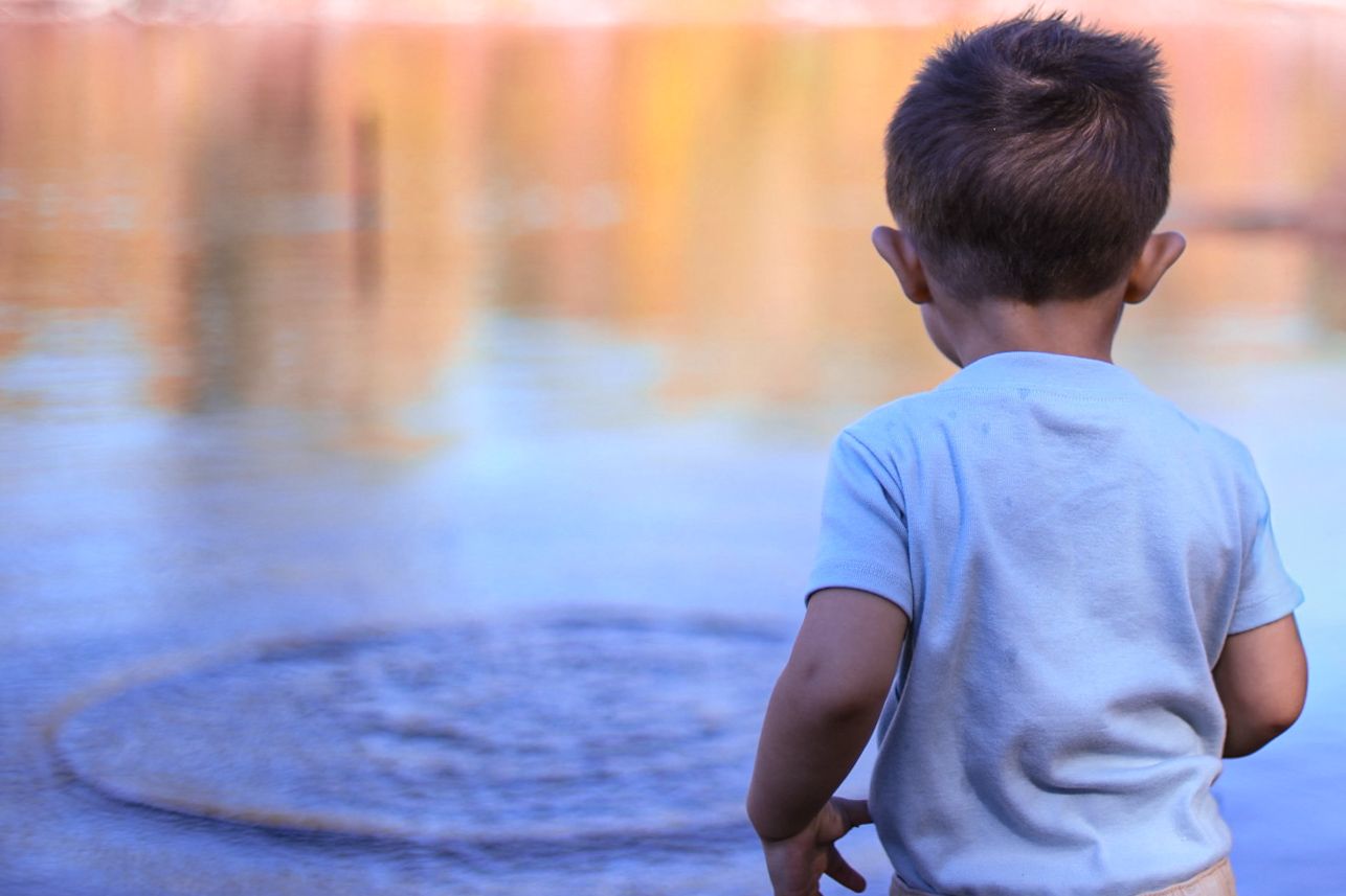 A toddler stands alone gazing at a calm reflective water surface on a sunny day.