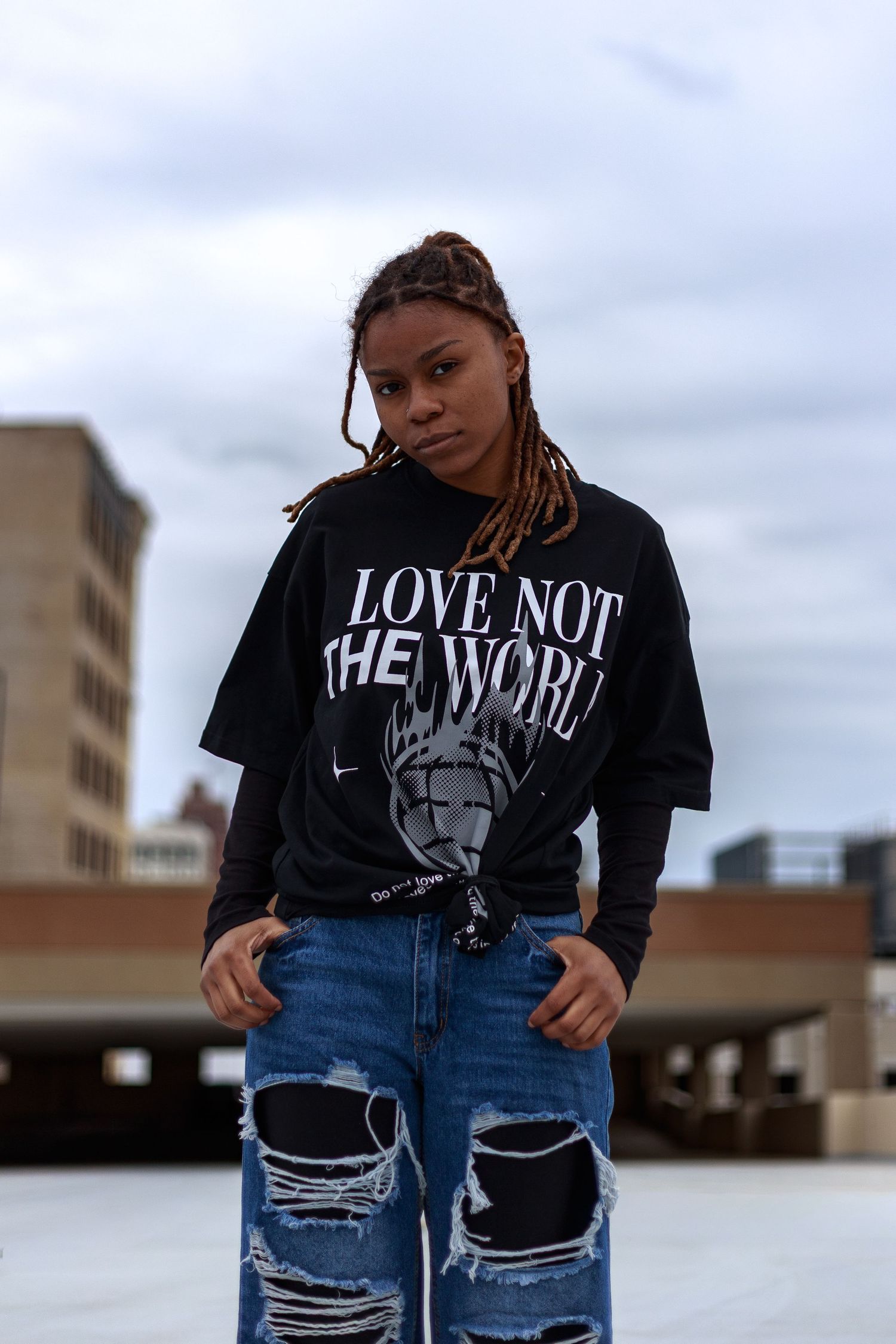 Young woman in ripped jeans and black Love Not The World tee poses confidently on an urban rooftop.