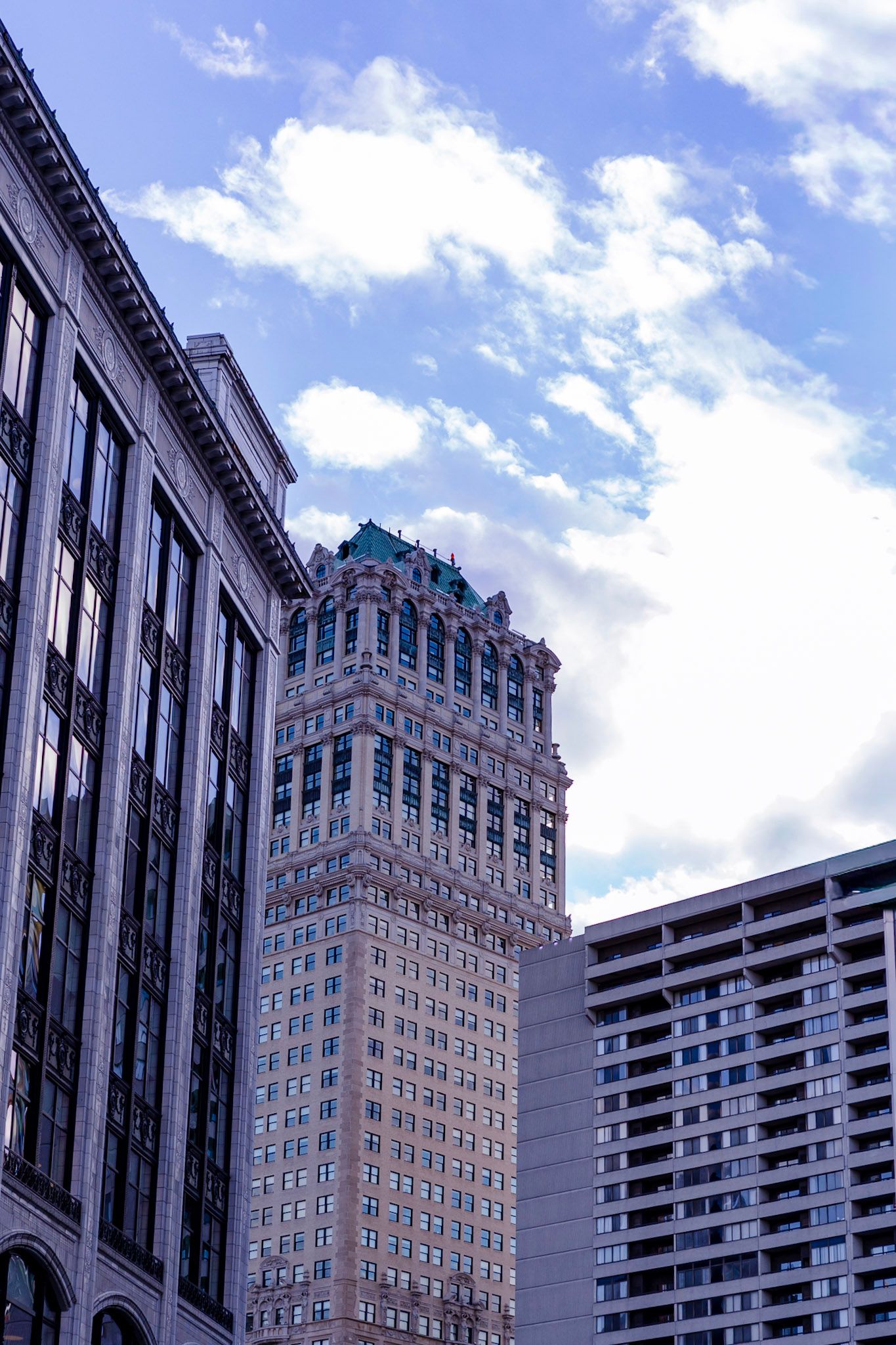 Historic ornate skyscraper with green crown detail rises between modern buildings under a partly cloudy sky.
