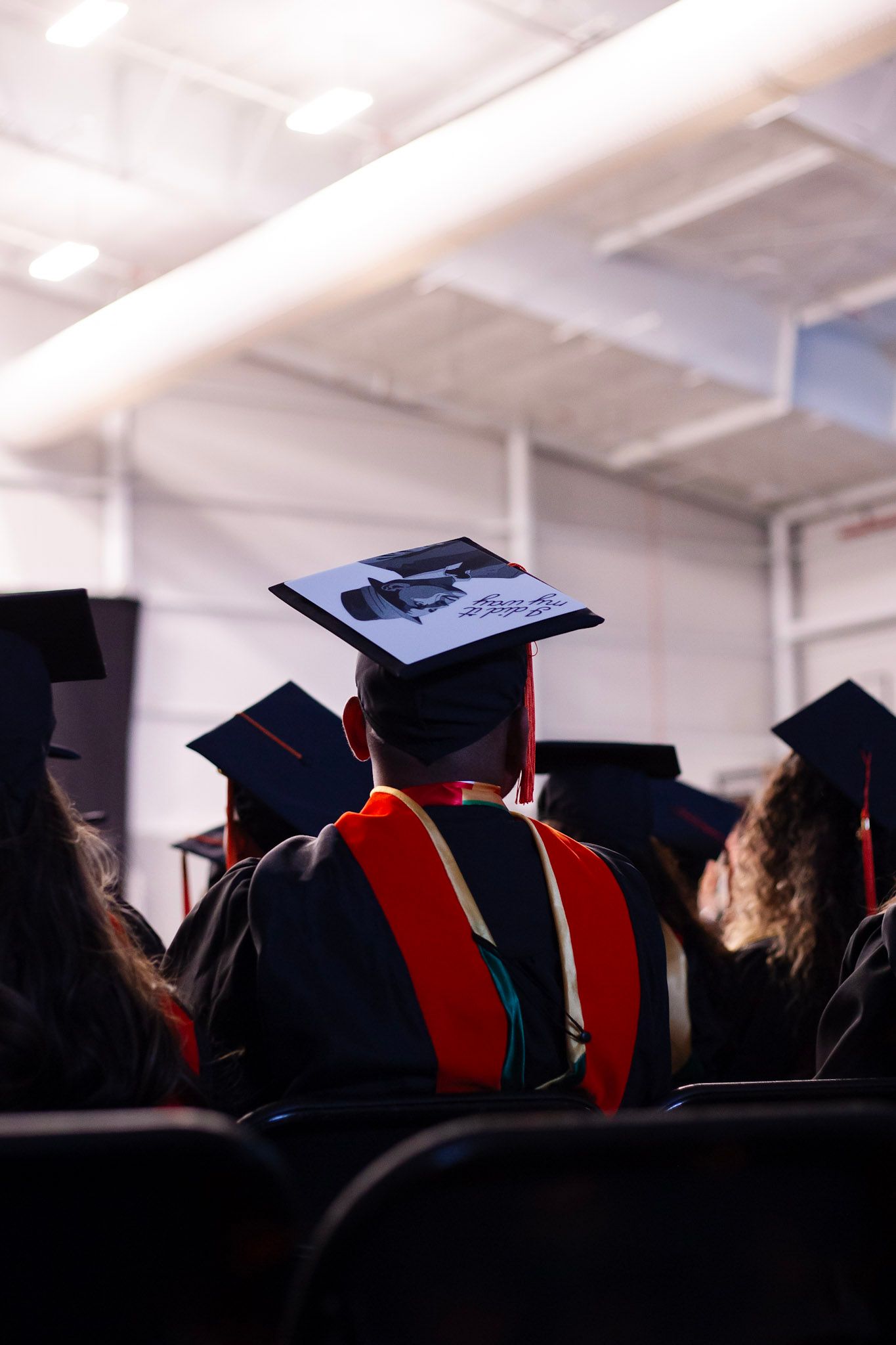 Graduate wearing decorated mortarboard cap with a photo and red hood seated among peers at commencement ceremony.