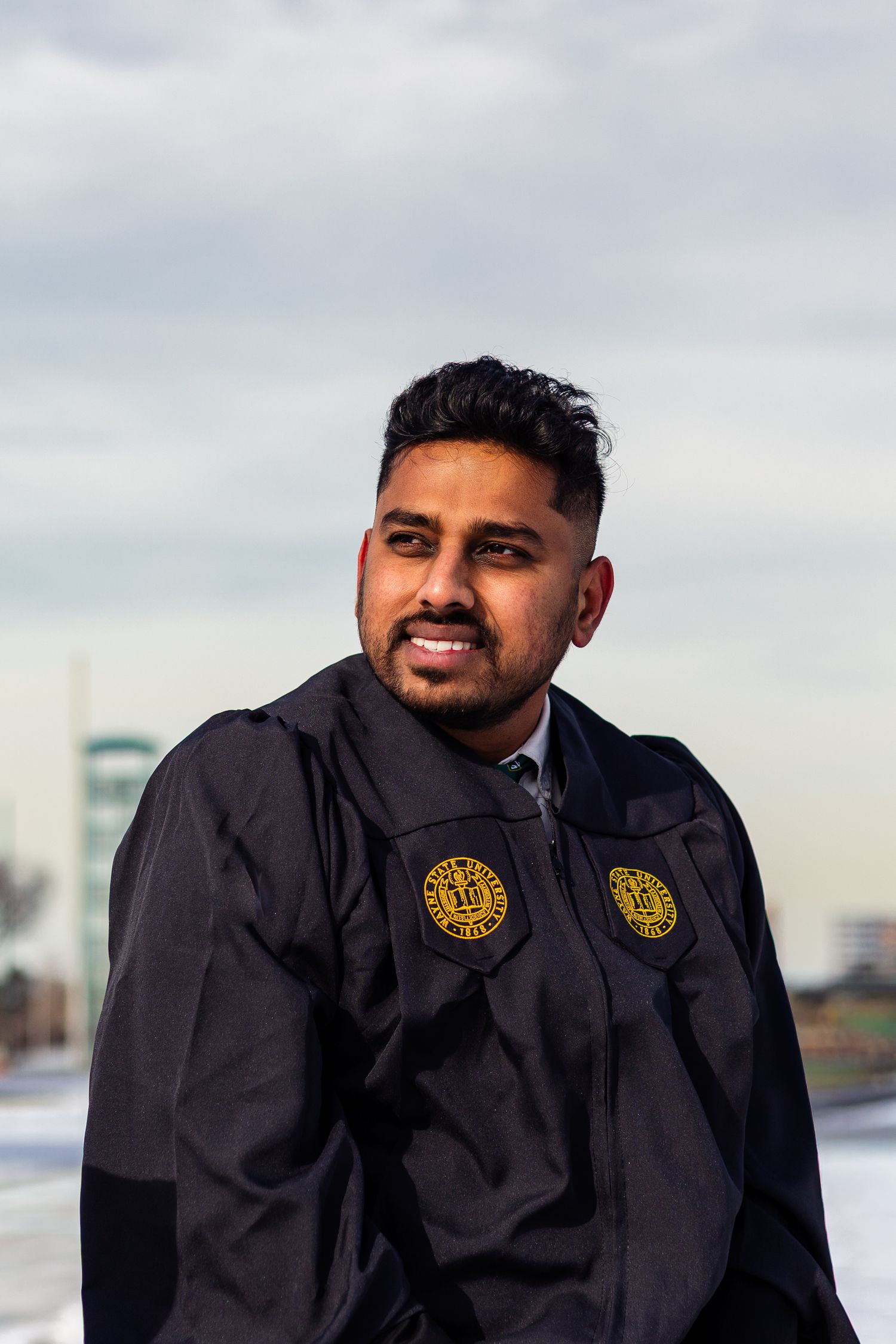 Young man in Wayne State University graduation gown smiles outdoors under cloudy sky.