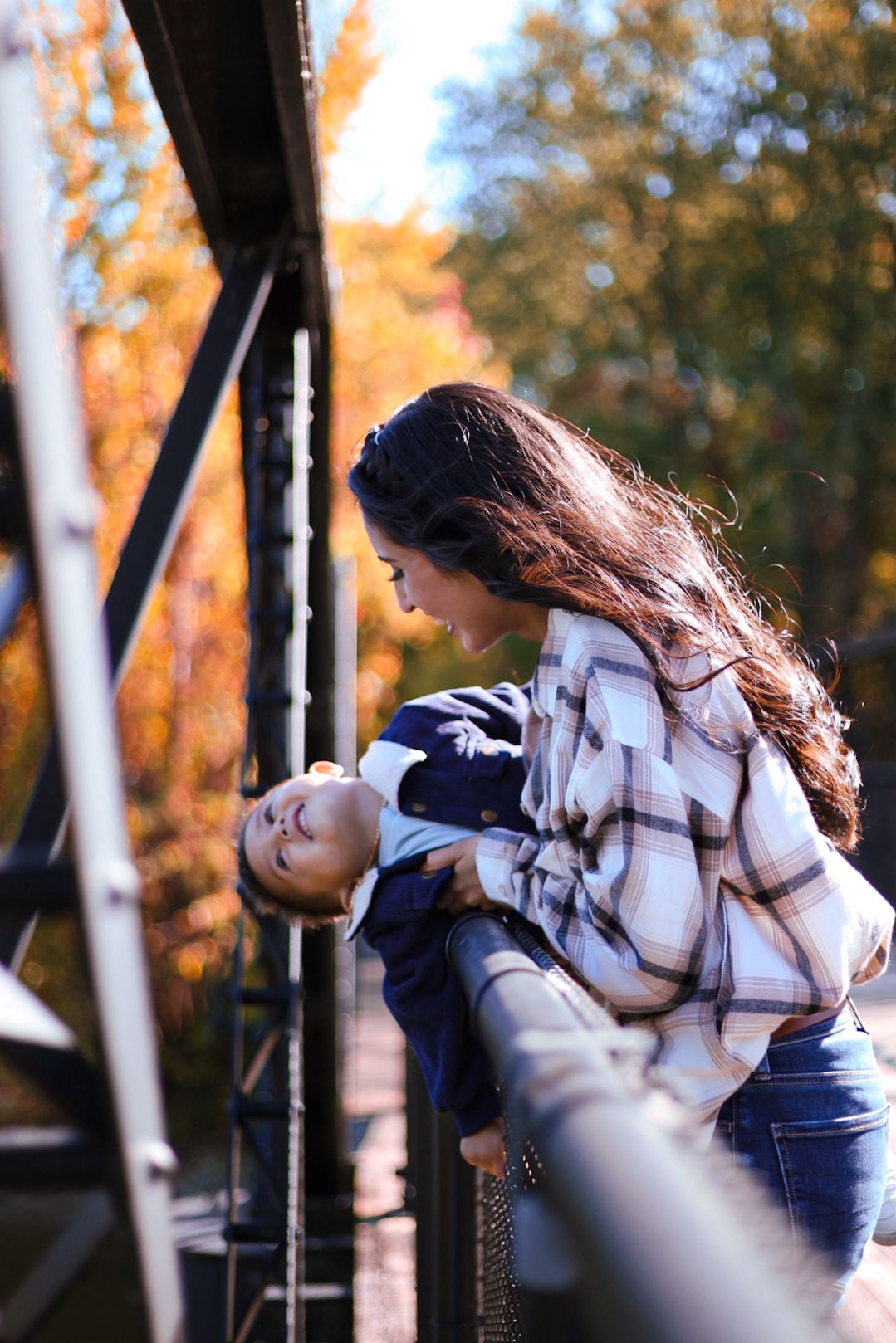 A mother playfully dips her baby backward on a bridge amid colorful autumn trees.
