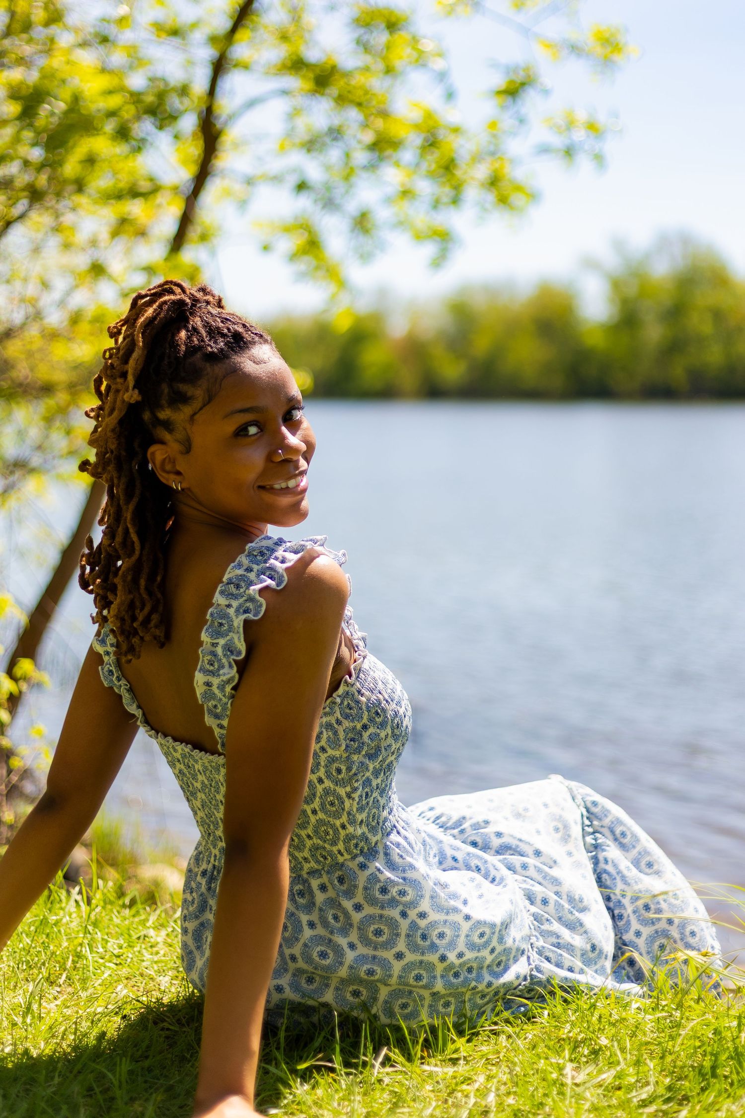 Young Black woman in floral sundress sitting on grass smiling by sunny lakeside.