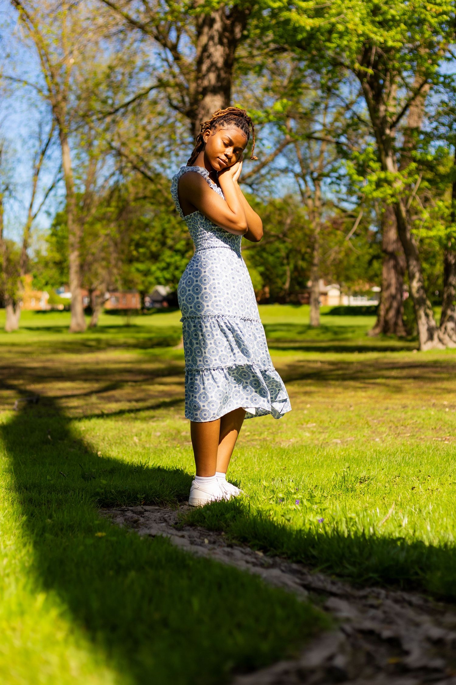 Young Black woman in blue floral sundress posing gracefully on a sunny park path with lush green grass.