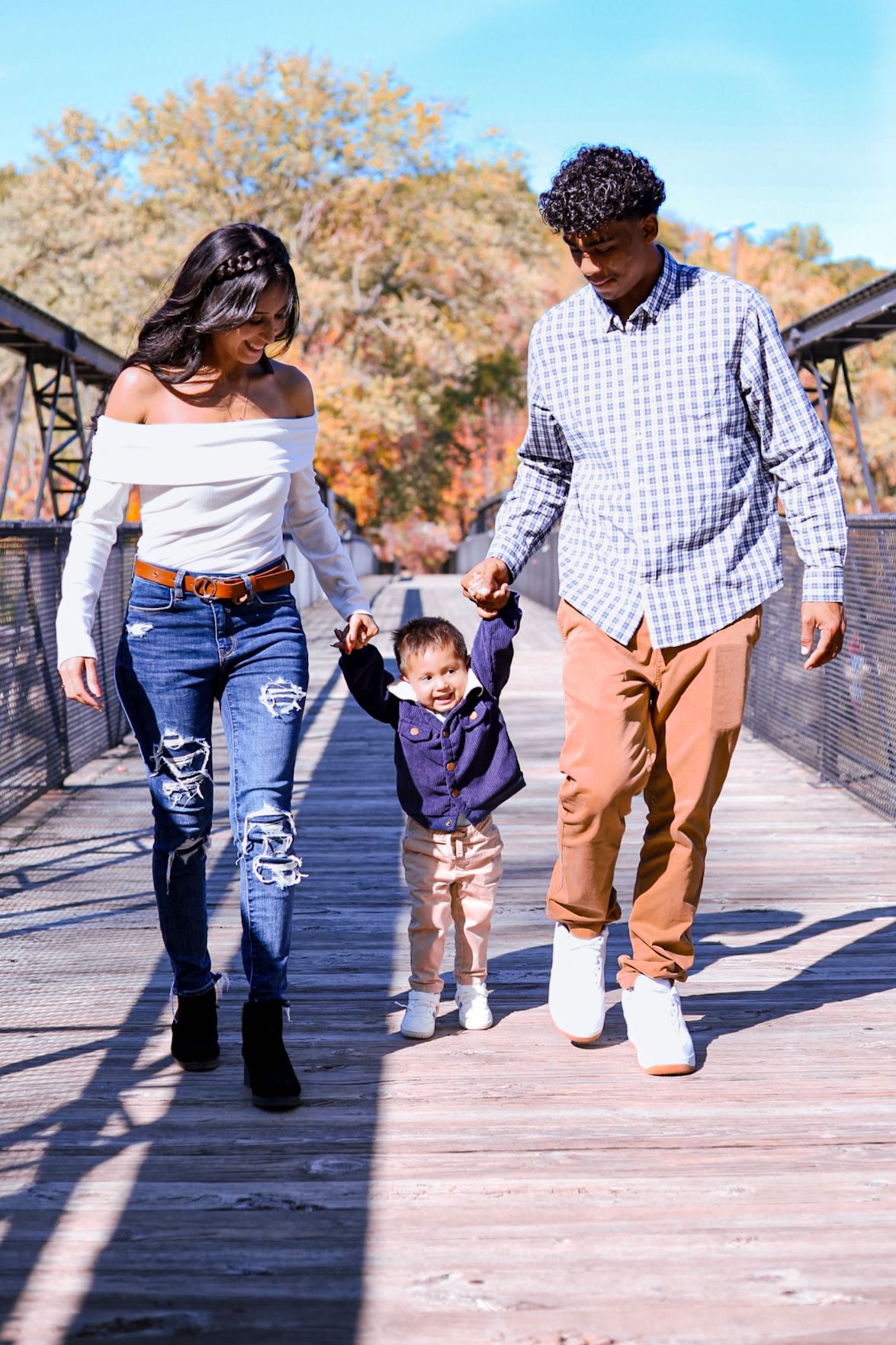 Happy young family with toddler walking hand-in-hand on wooden bridge during autumn day.