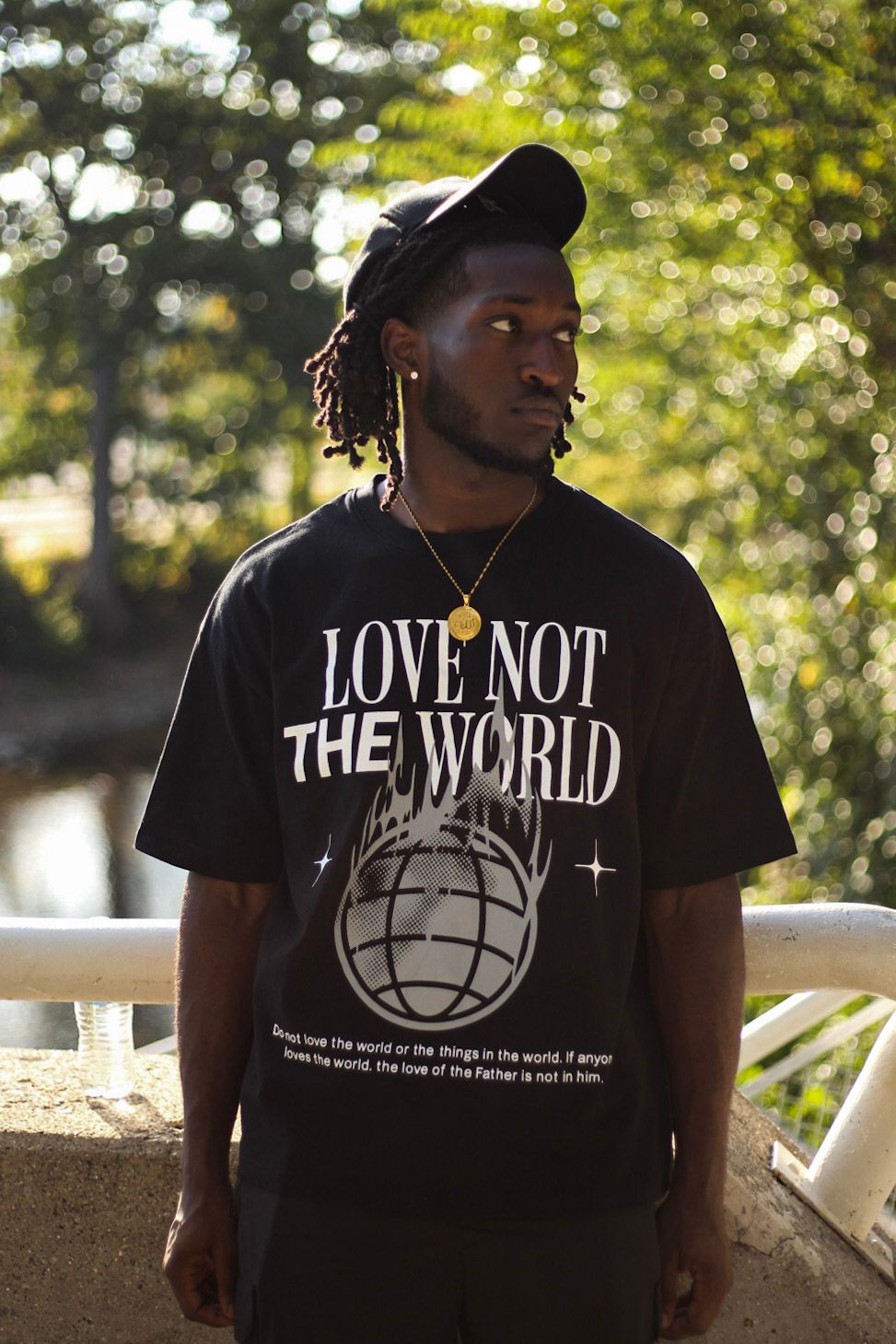 Man in black 'Love Not The World' tee with gold chain and black cap standing on bridge outdoors.