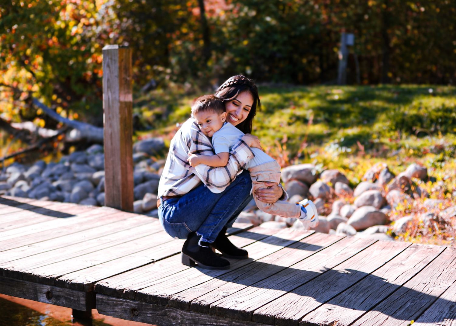 A mother crouches on a wooden dock hugging her toddler son surrounded by fall foliage.