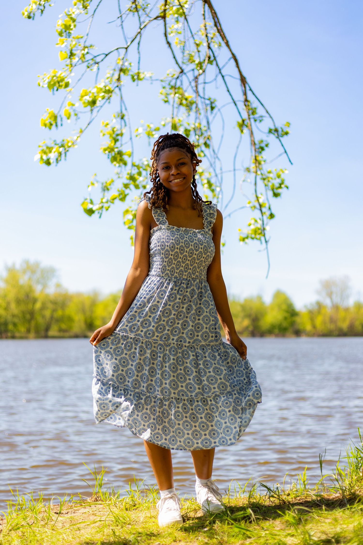 Young Black woman twirling in a blue floral sundress by a lakeside under blooming spring tree branches.