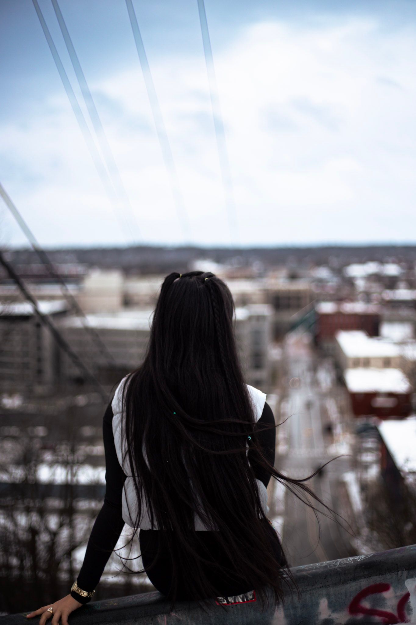 Woman with long dark hair sitting on railing overlooking snowy urban cityscape from above.